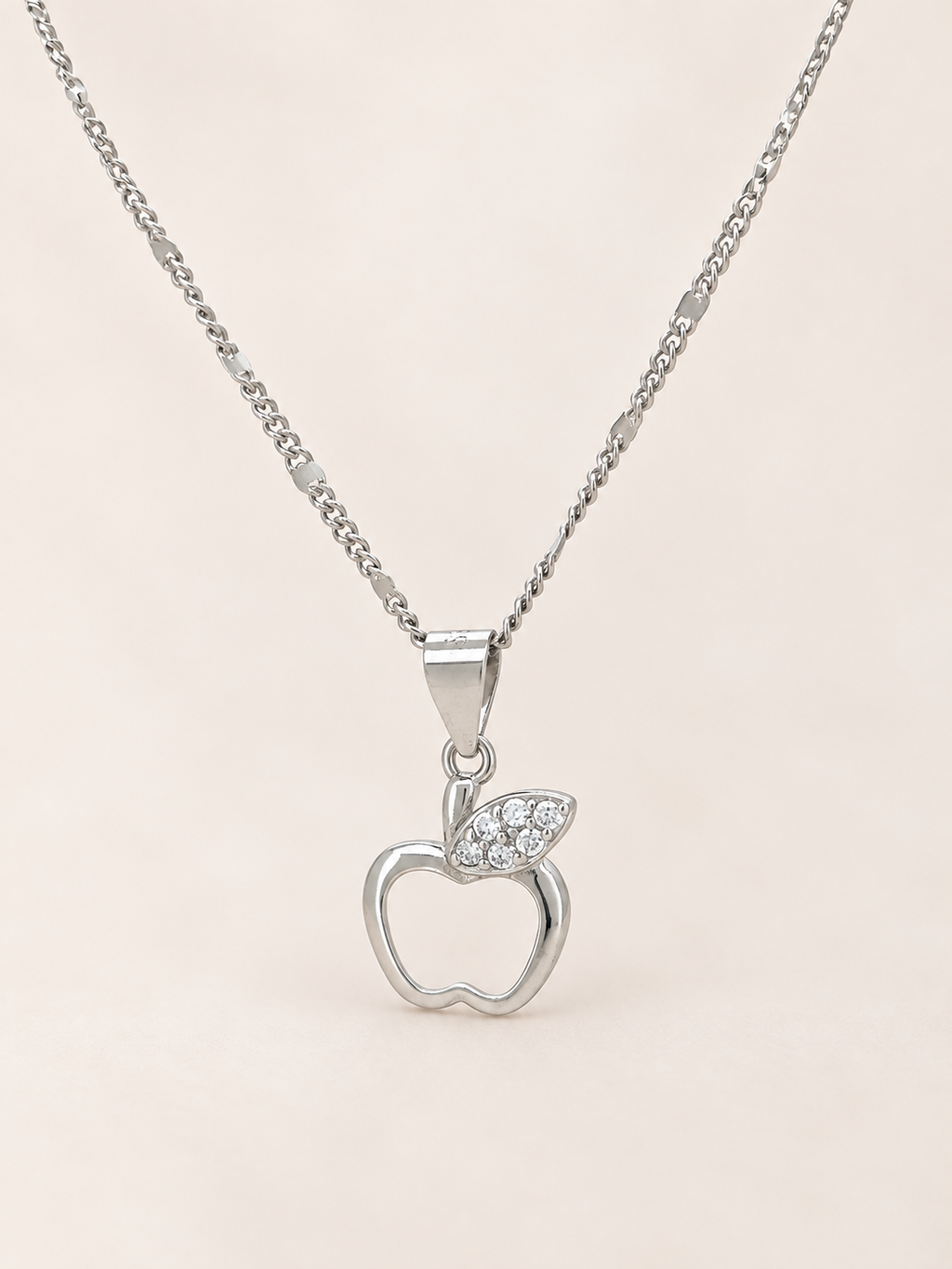 A silver apple pendant necklace with a small leaf detail paved with clear stones, shown on a delicate silver chain against a soft beige background.