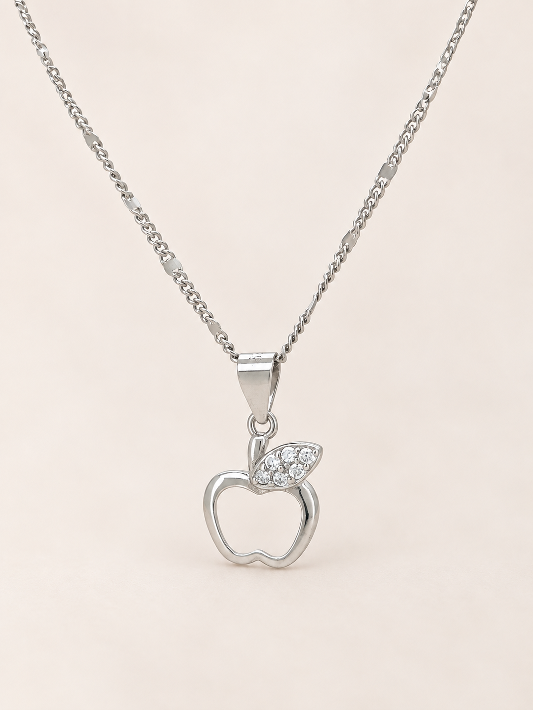 A silver apple pendant necklace with a small leaf detail paved with clear stones, shown on a delicate silver chain against a soft beige background.