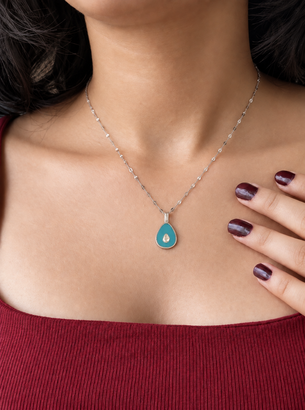A silver teardrop pendant necklace featuring a vibrant turquoise blue enamel body with a small teardrop in the center, suspended from a silver faceted chain worn by a model wearing a red top.