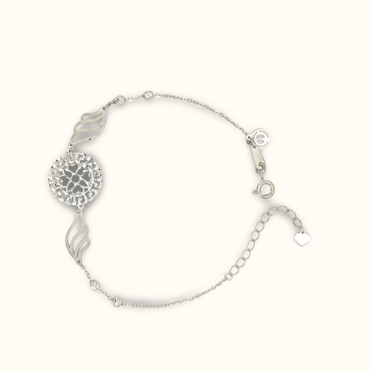 A silver-toned delicate chain bracelet featuring a textured circular medallion, two stylized leaf charms, and a small heart charm, displayed on a clean off-white background.