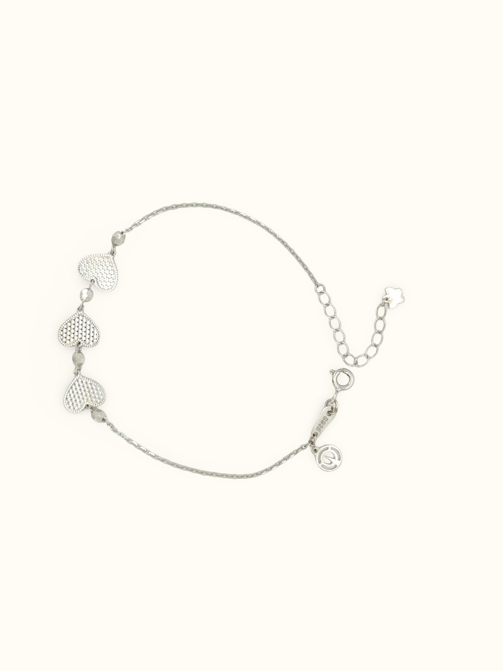 A delicate silver chain bracelet featuring three textured heart charms and a small heart-shaped tag, displayed on a solid off-white background.