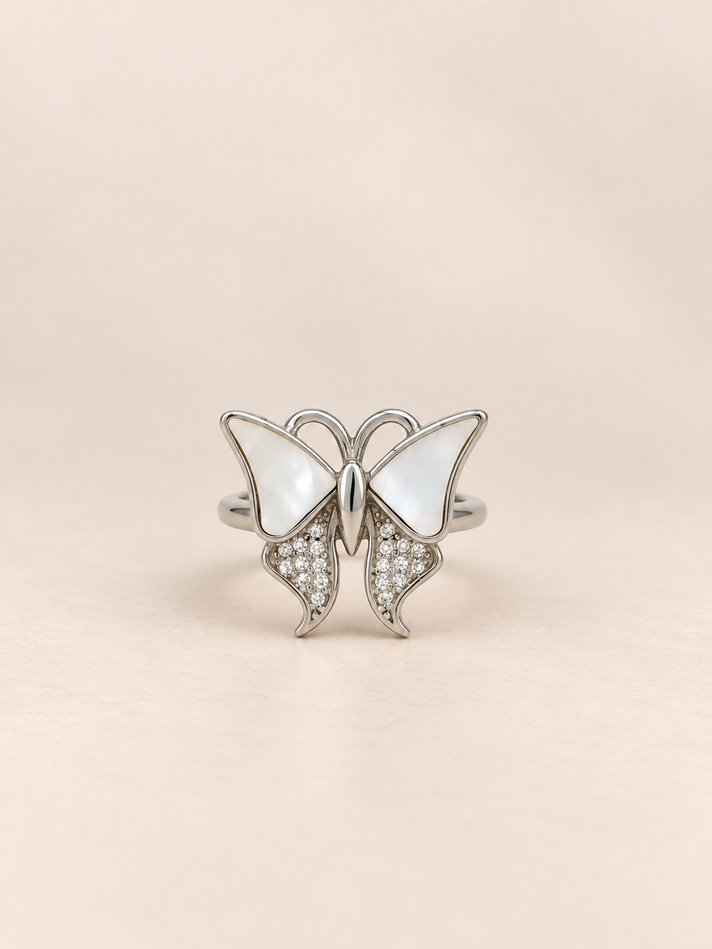 A silver butterfly ring featuring mother-of-pearl wings and pavé-set clear crystals on the lower wings, displayed on a soft beige background.
