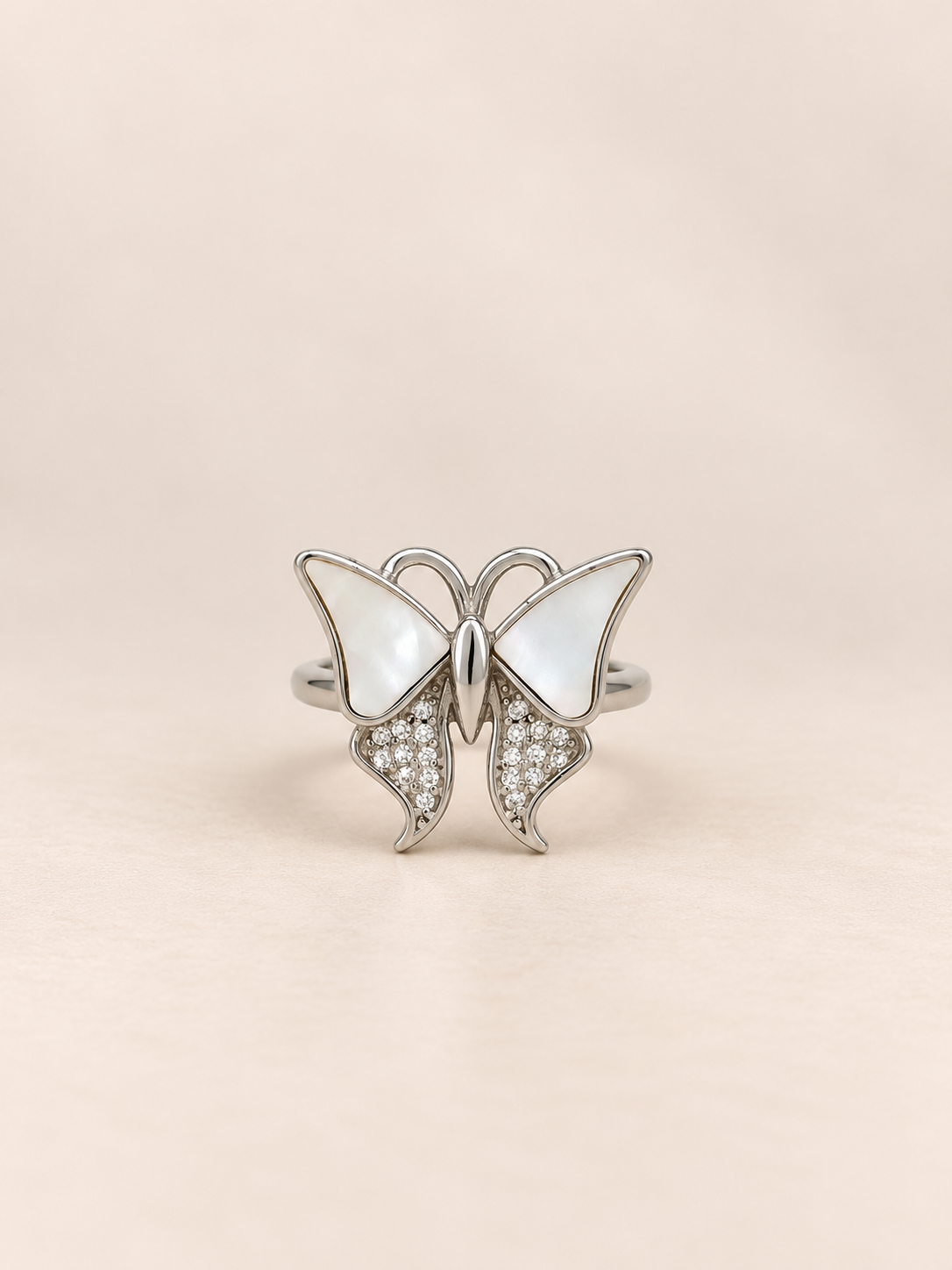 A silver butterfly ring featuring mother-of-pearl wings and pavé-set clear crystals on the lower wings, displayed on a soft beige background.