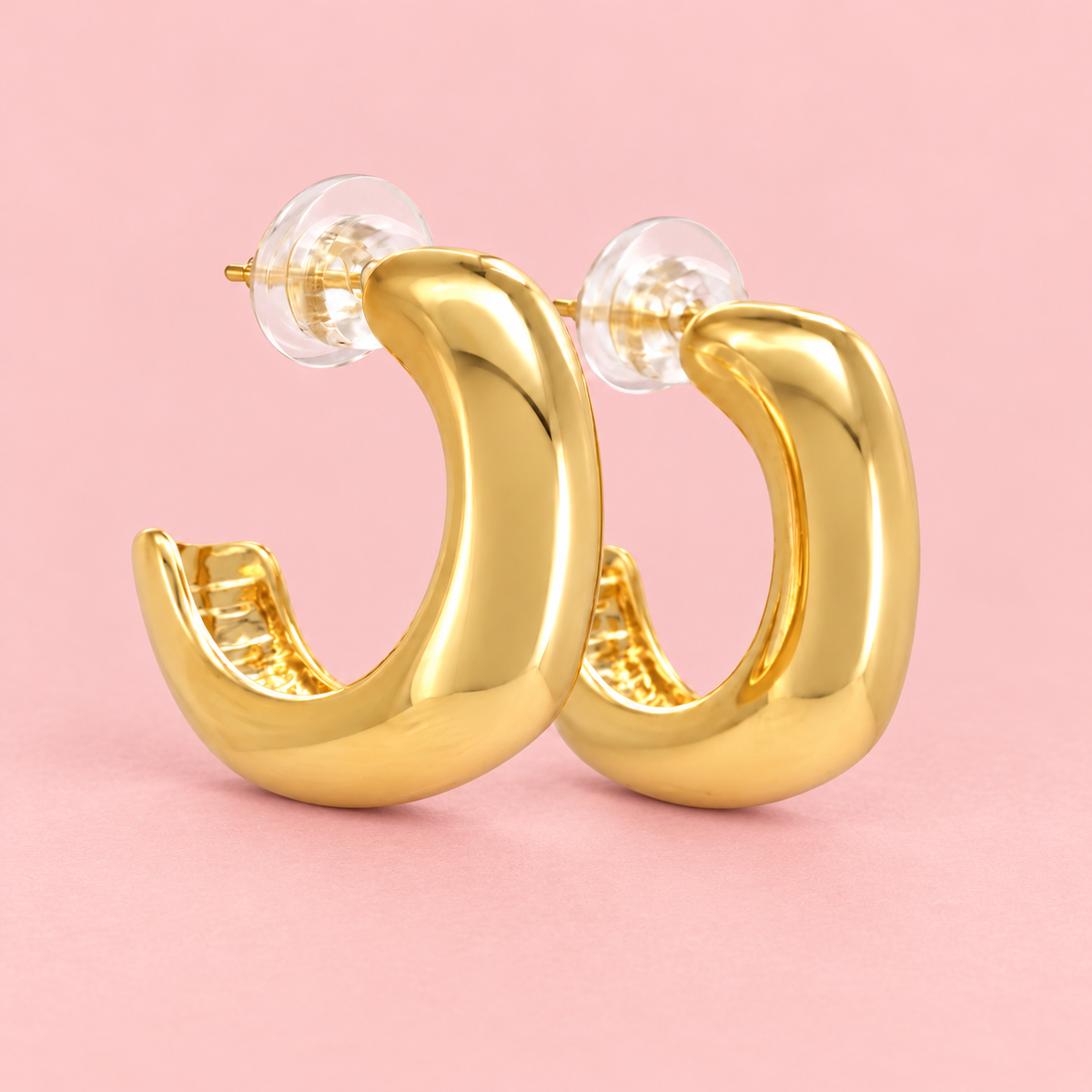Gold-tone chunky huggie hoop earrings with a smooth, rounded shape and polished finish displayed against a pink background.