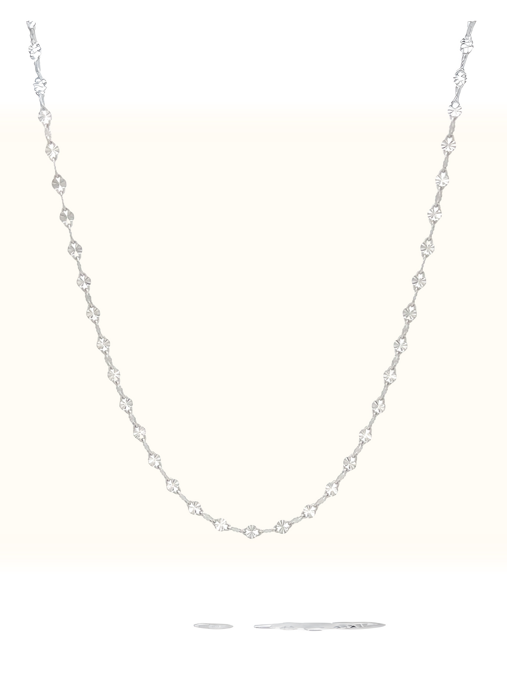 A delicate silver-toned tennis necklace featuring small, clear round-cut gemstones set along a fine chain, displayed against a solid off-white background.