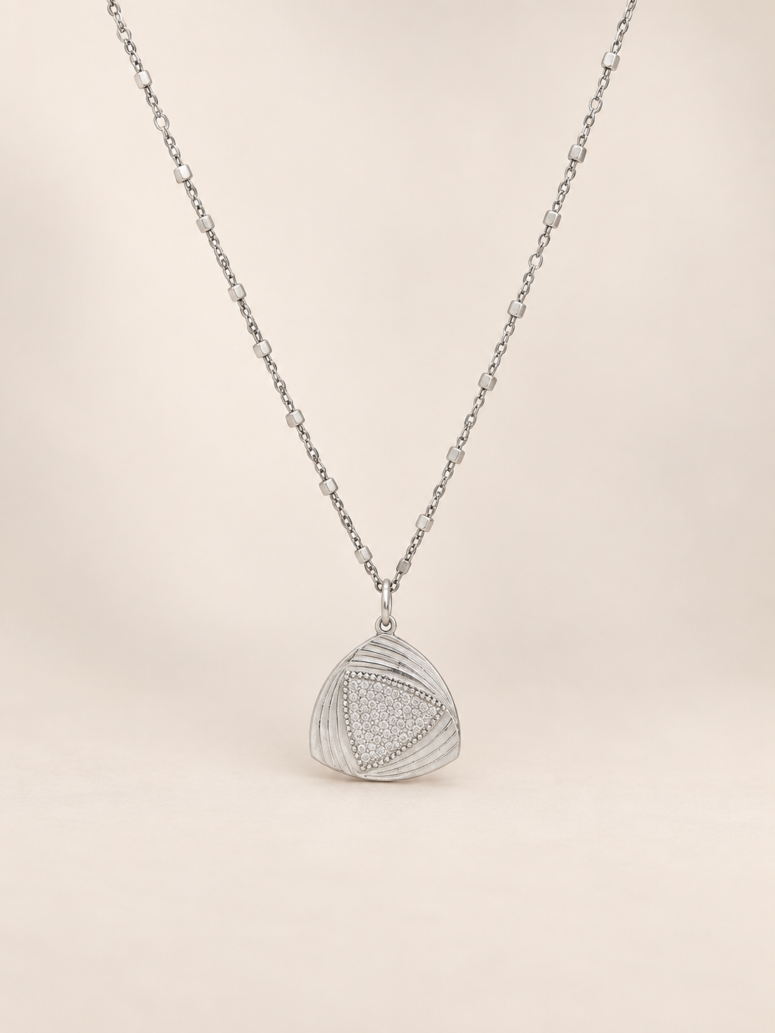A silver pendant necklace featuring a textured, geometric triangular design with a pavé-style inset, suspended from a delicate silver chain against a solid, warm off-white background.