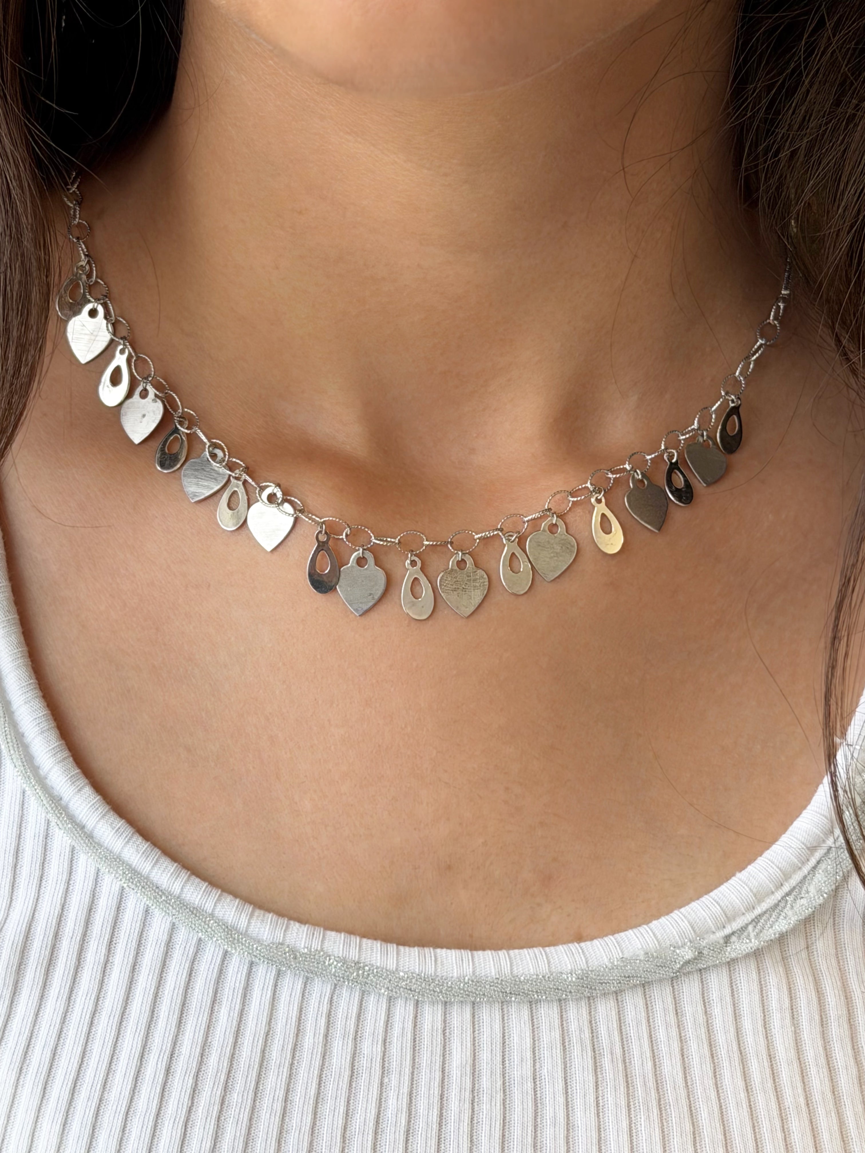 A close-up of a model's neck wearing a delicate silver-toned charm necklace with multiple small heart and teardrop-shaped pendants, paired with a white ribbed top.