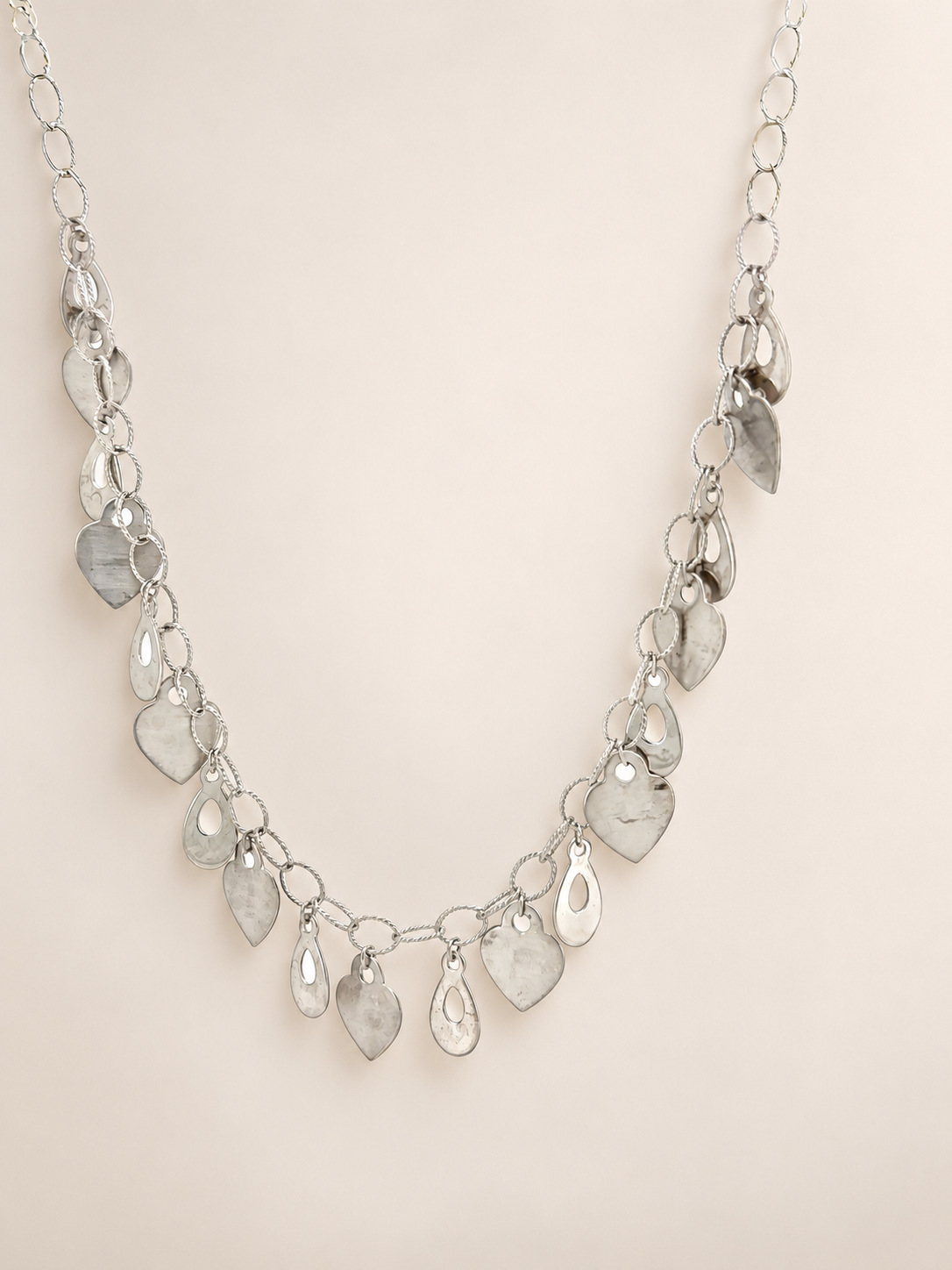 A silver-toned statement necklace featuring multiple dangling teardrop and leaf-shaped clear faceted stones on a delicate chain, displayed against a solid light beige background.
