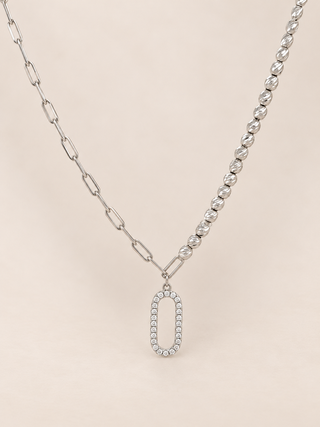 A silver-toned necklace featuring a dual-chain design with a paperclip chain on one side and a beaded chain on the other, both leading to an oval pendant encrusted with clear stones, set against a solid pale pink background.
