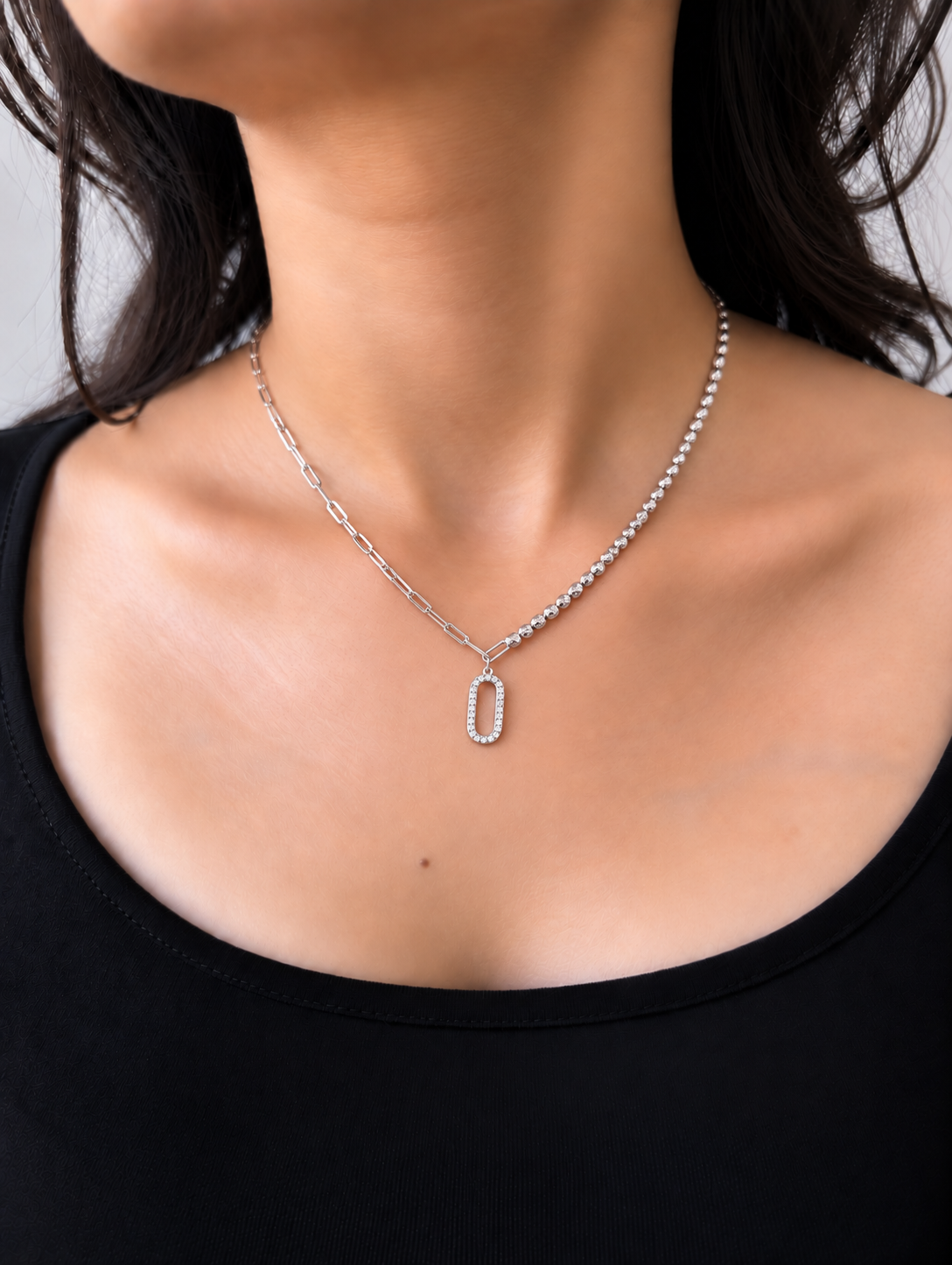 A silver-toned necklace featuring a dual-chain design with a paperclip chain on one side and a beaded chain on the other, both leading to an oval pendant encrusted with clear stones, worn by a model.