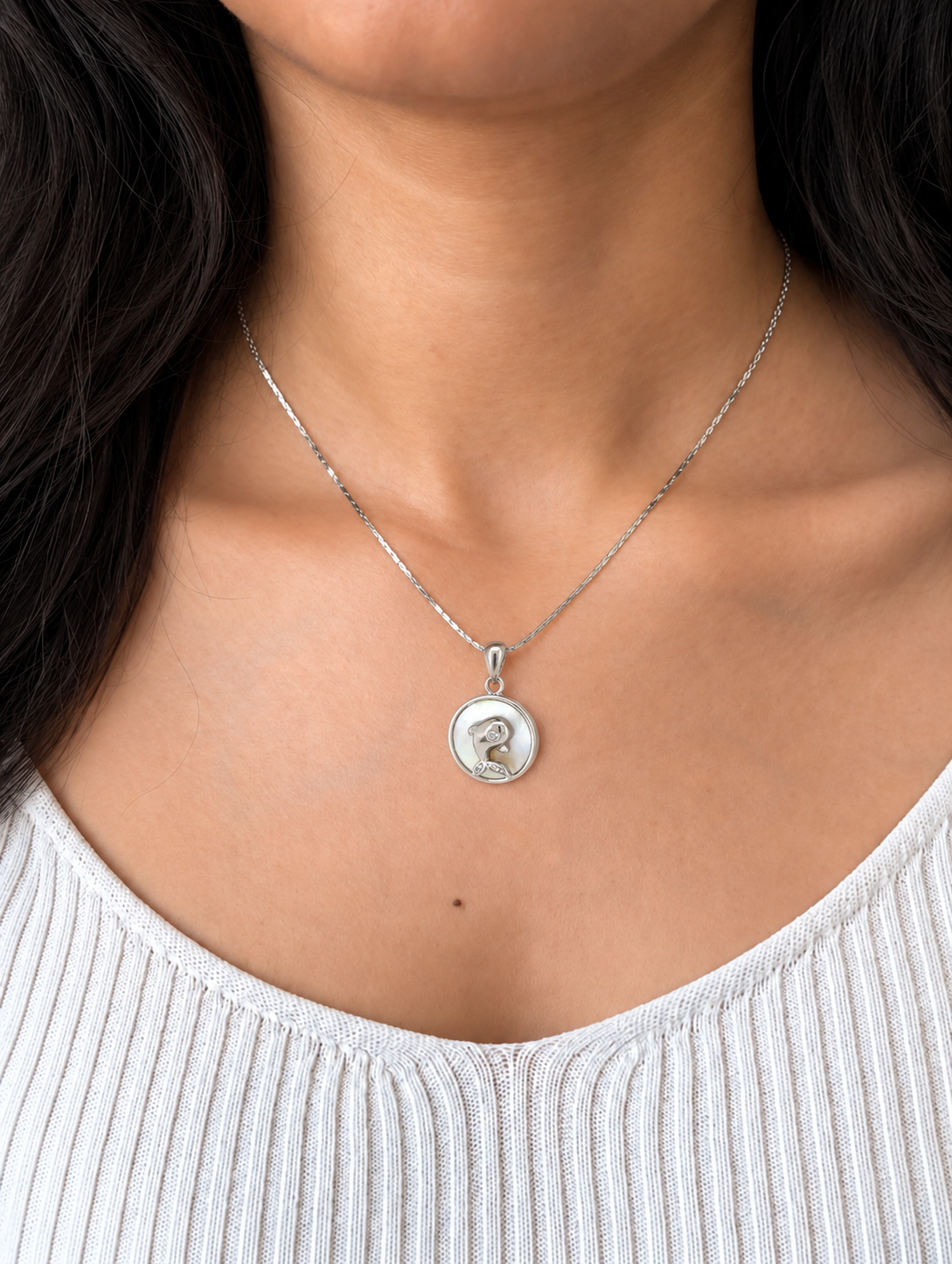 A silver pendant necklace featuring a circular charm with a polished silver dog silhouette and a small clear gemstone, suspended from a delicate silver chain which is worn by a model wearing a white top.