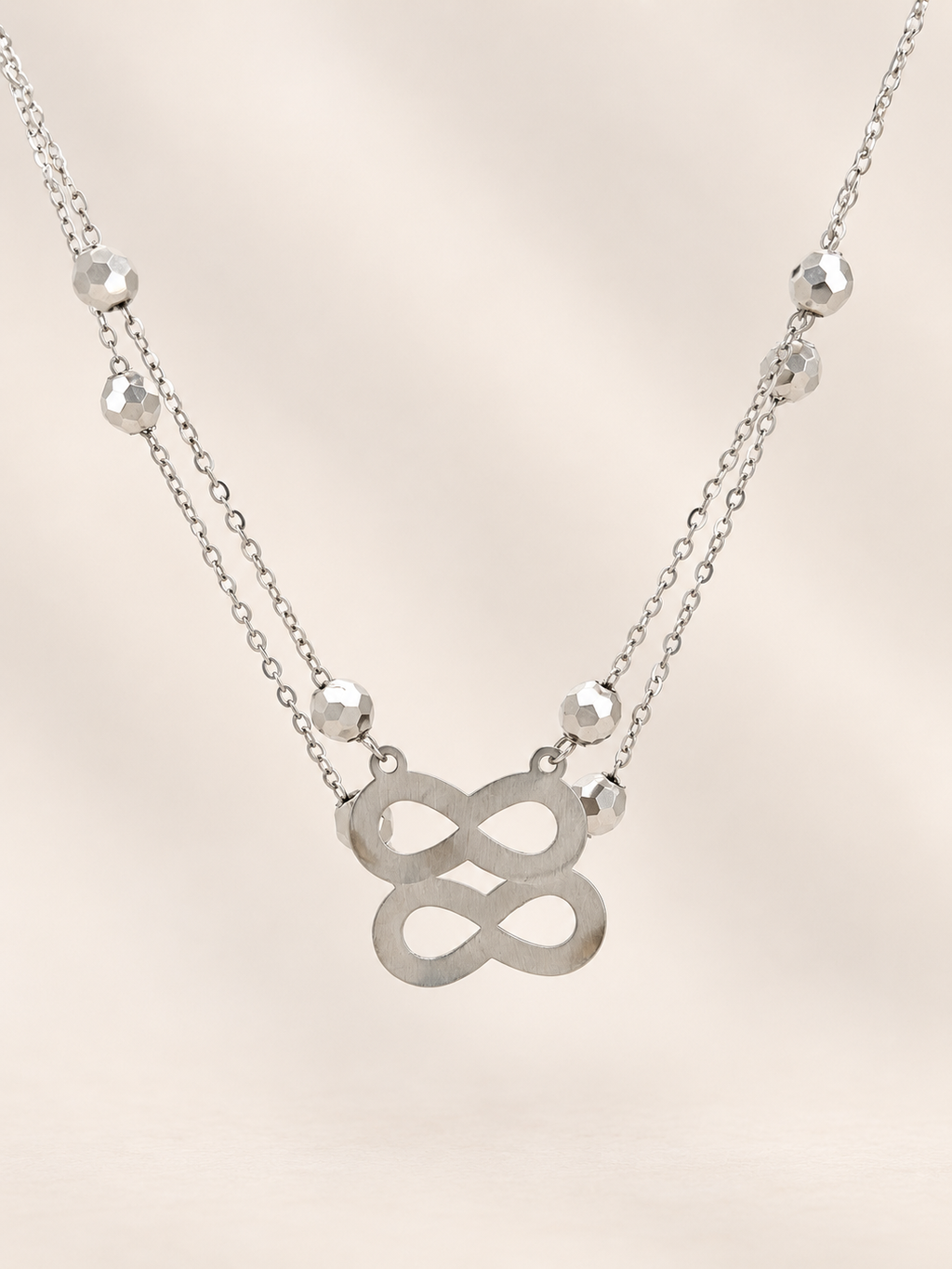 A silver double-layered necklace featuring a central infinity symbol pendant and small faceted bead accents on a delicate chain, set against a soft beige background.