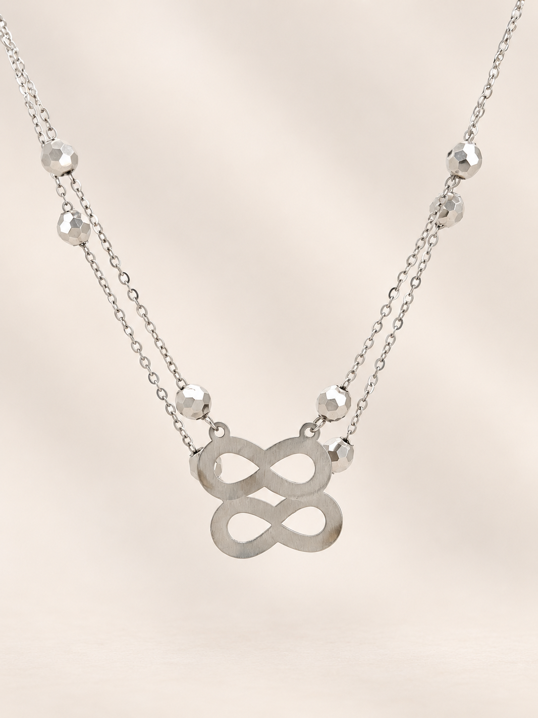 A silver double-layered necklace featuring a central infinity symbol pendant and small faceted bead accents on a delicate chain, set against a soft beige background.