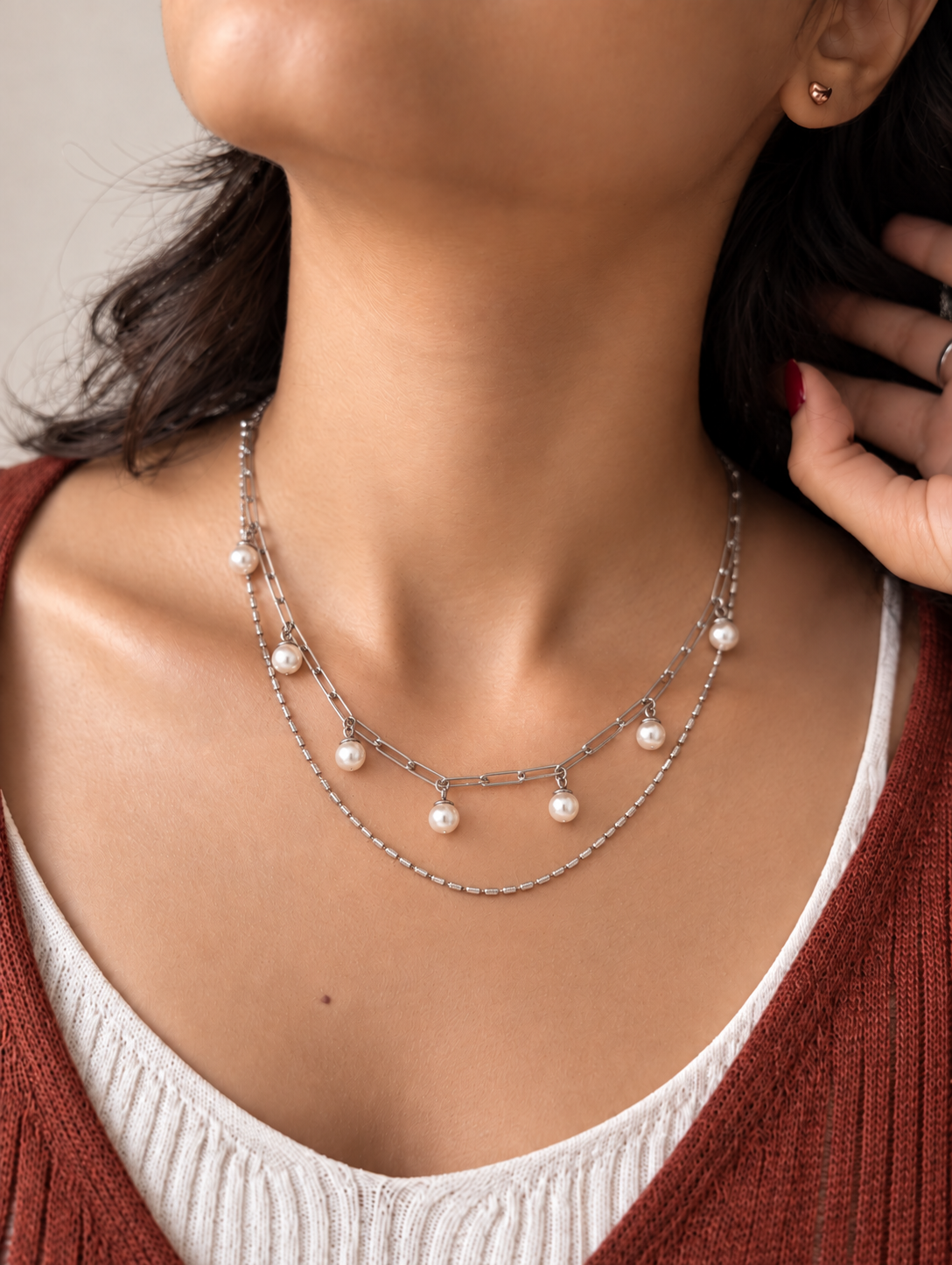 A close-up of a model wearing a double-layered silver chain necklace with small white pearl beads, styled with a white ribbed tank top and a rust-colored knit cardigan.