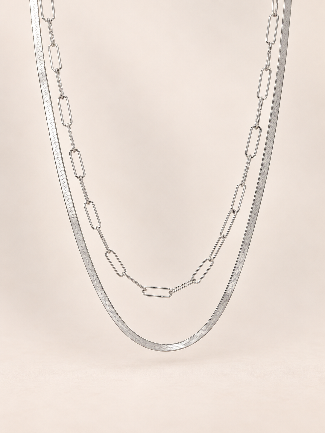 A silver paperclip chain necklace draped in a U-shape against a solid, warm beige background.