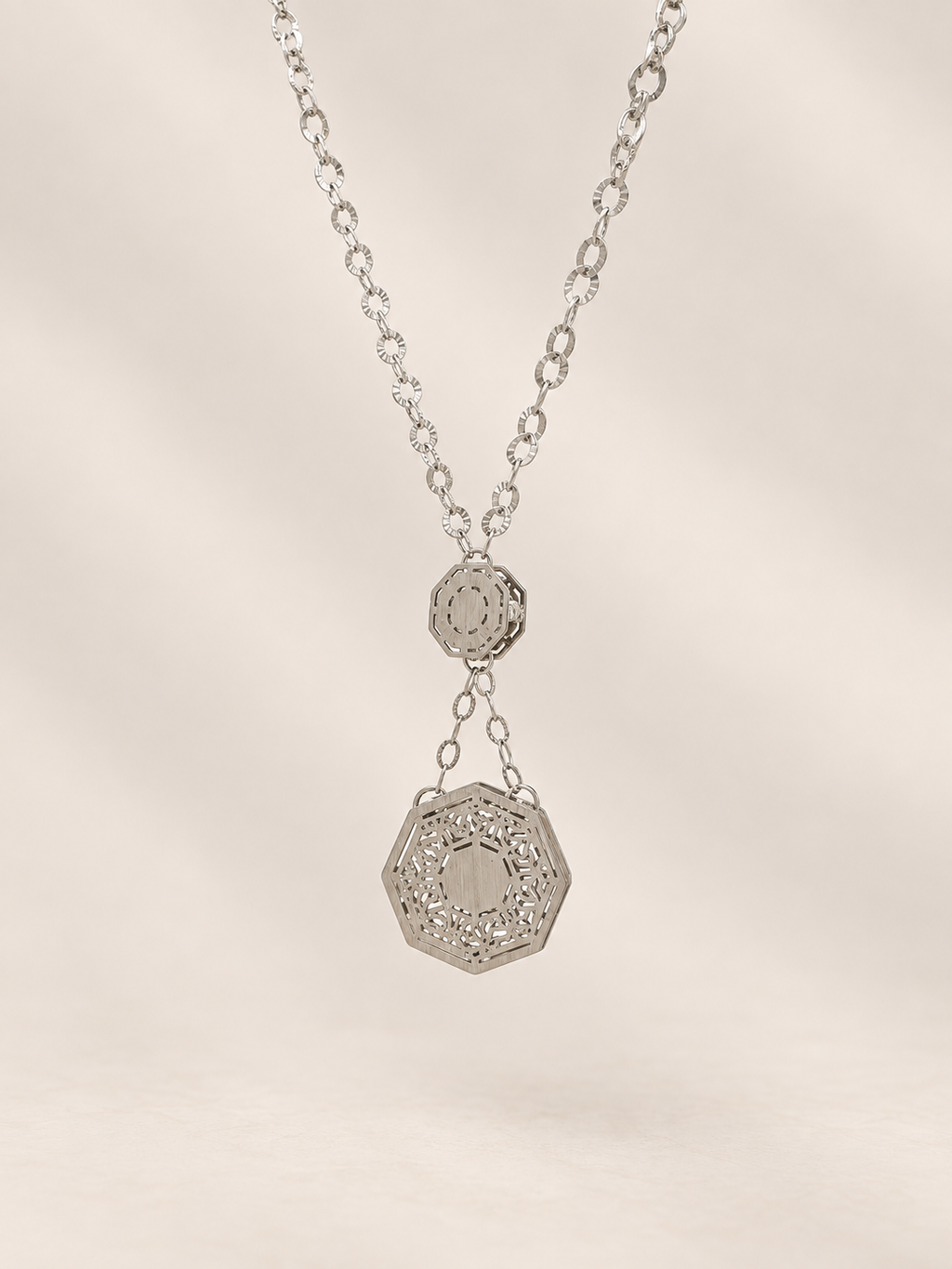 A silver-toned necklace featuring a delicate chain and a two-tiered pendant design, with a larger octagonal pendant hanging below a smaller circular one, set against a soft beige background.