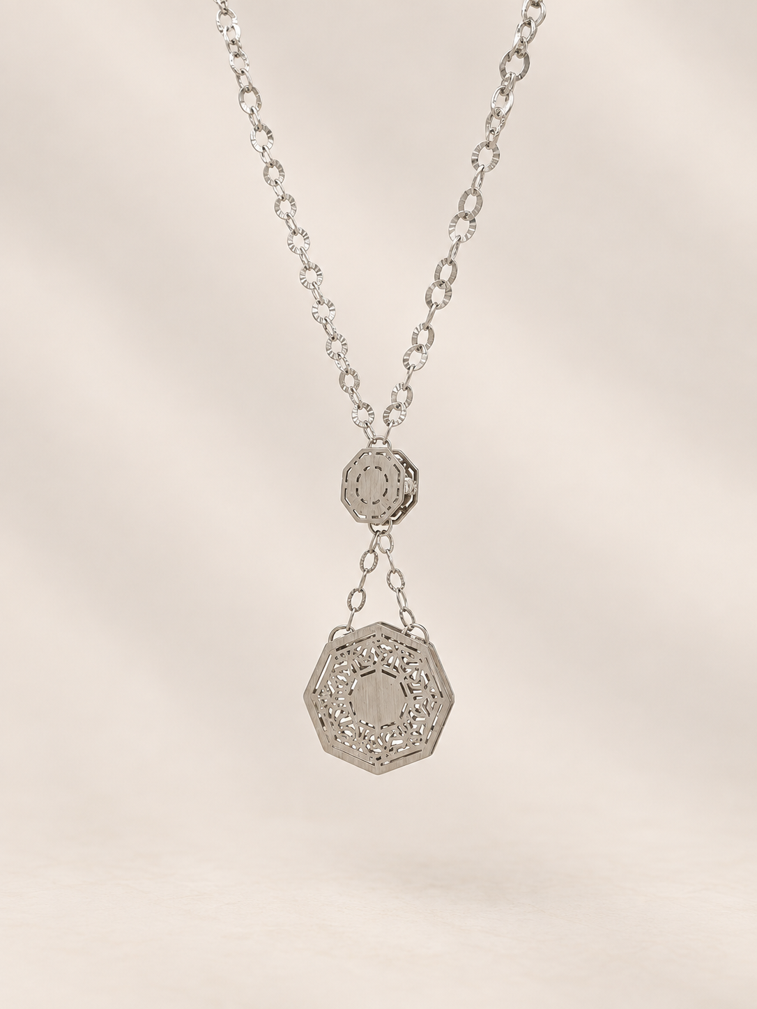 A silver-toned necklace featuring a delicate chain and a two-tiered pendant design, with a larger octagonal pendant hanging below a smaller circular one, set against a soft beige background.