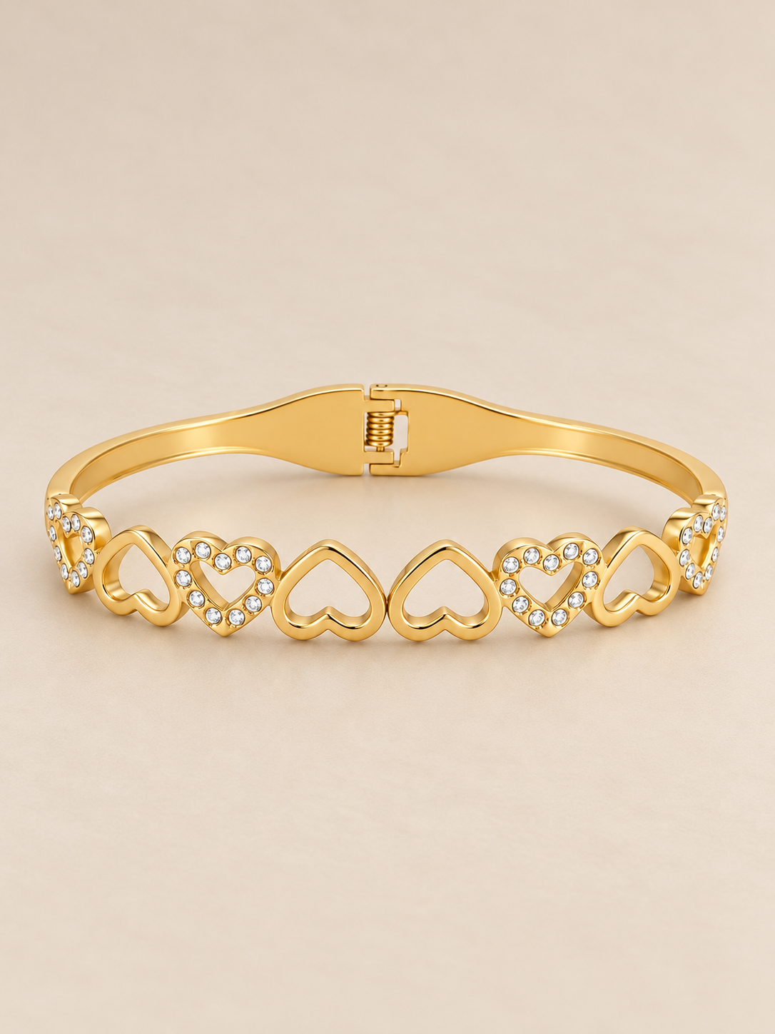Gold-toned stainless steel bangle featuring alternating open-heart motifs with polished finishes and sparkling white stone accents with elegant hinged cuff design, displayed on a clean pink background.