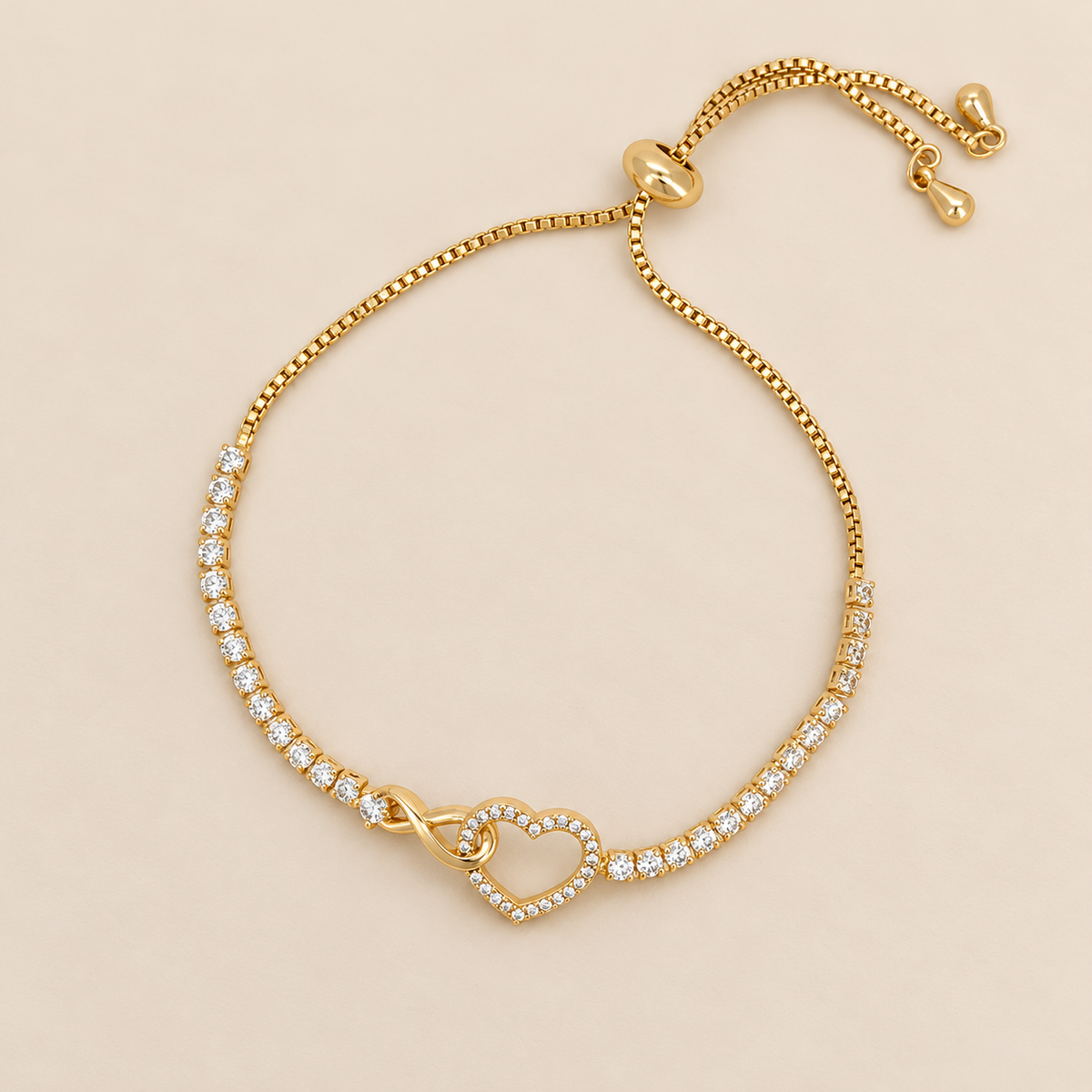 Gold-toned stainless steel bracelet featuring a sparkling open heart intertwined with an infinity motif and white stone accents, adjustable slider chain bracelet with a polished golden finish, displayed on a clean pink background.