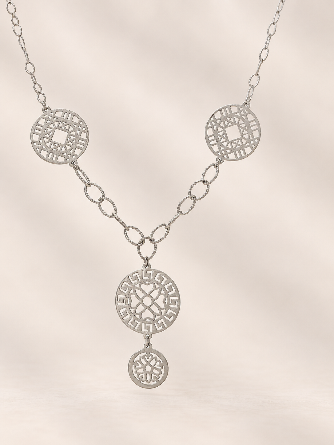 A silver-toned necklace featuring three circular filigree medallions connected by delicate chain links, with a larger central pendant and a smaller dangling charm, shown against a soft beige background.