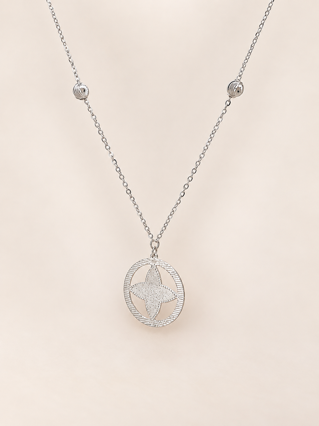 A silver-toned necklace featuring a delicate chain with small bead accents and a central oval pendant containing a textured star design, displayed against a soft, light beige background.