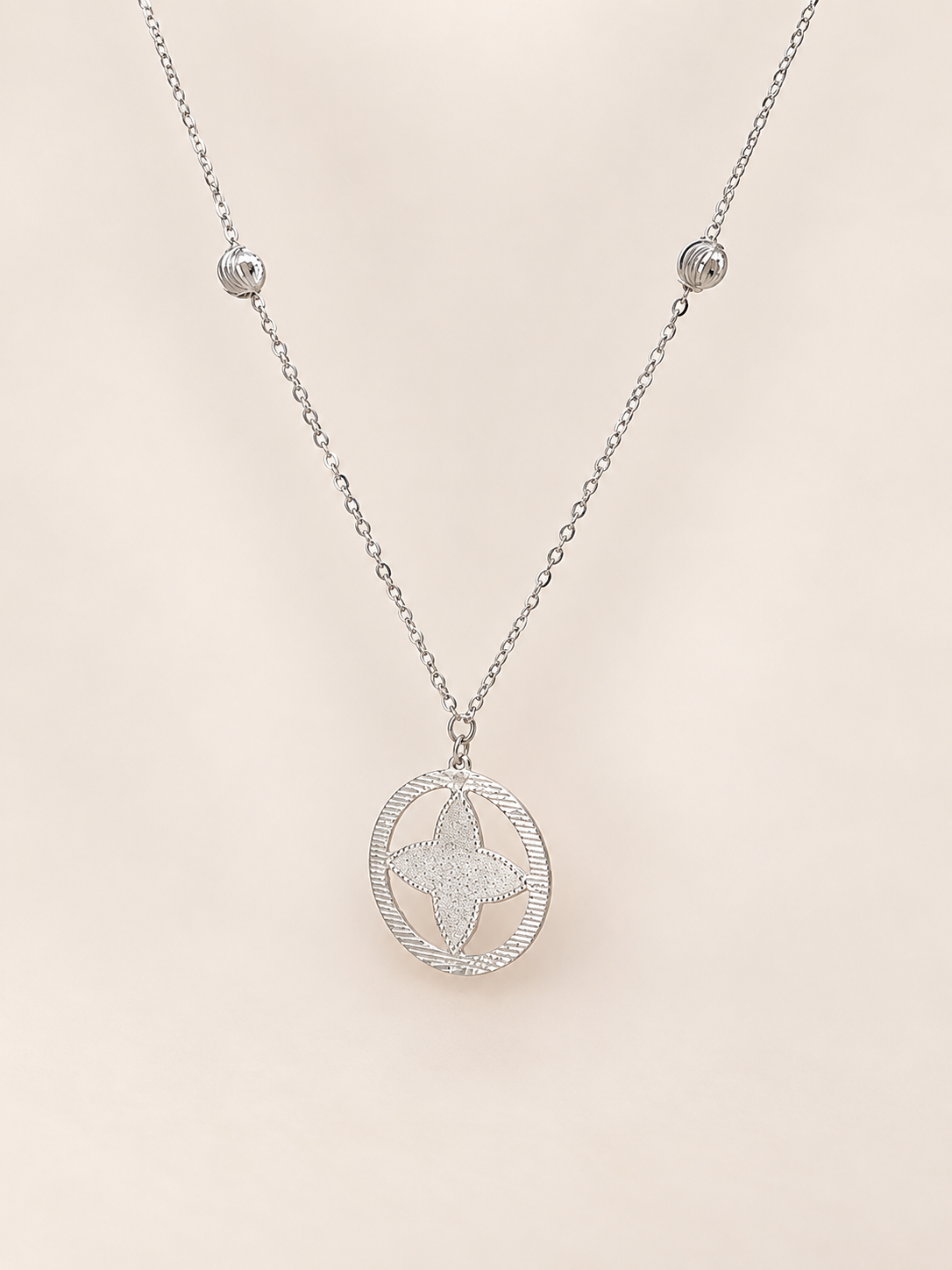 A silver-toned necklace featuring a delicate chain with small bead accents and a central oval pendant containing a textured star design, displayed against a soft, light beige background.