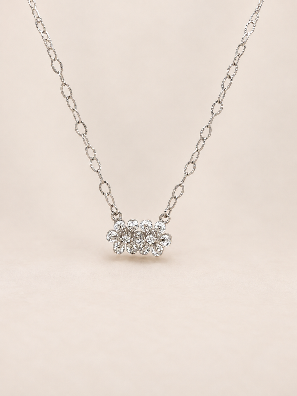A silver-toned chain necklace featuring a small cluster of clear gemstones in a flower setting, displayed against a soft beige background.
