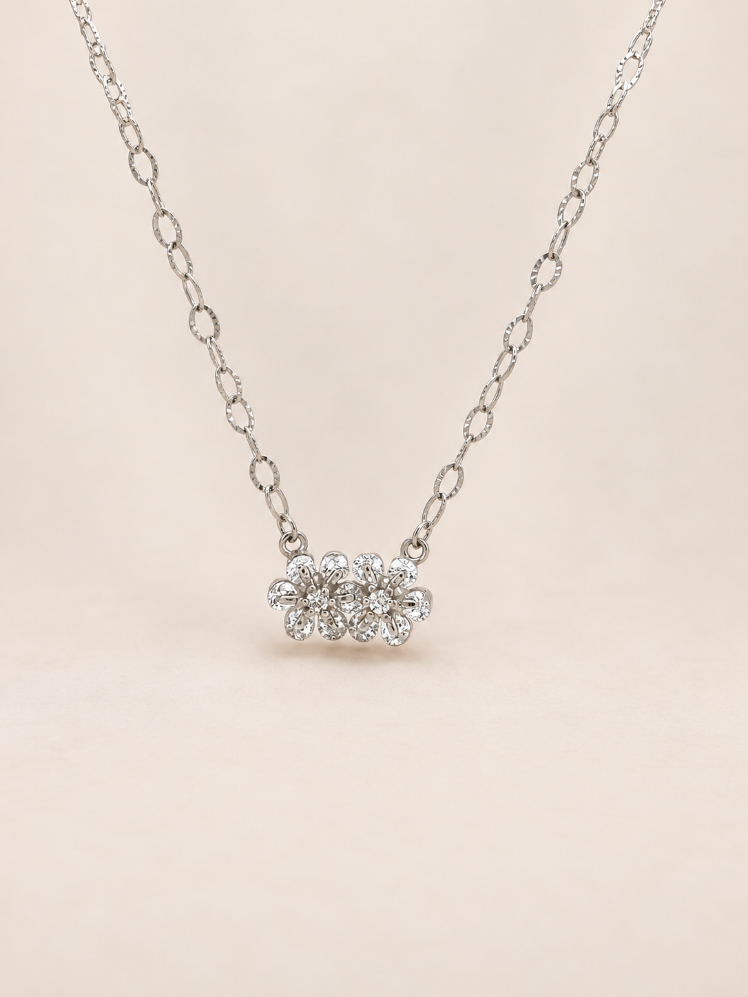 A silver-toned chain necklace featuring a small cluster of clear gemstones in a flower setting, displayed against a soft beige background.