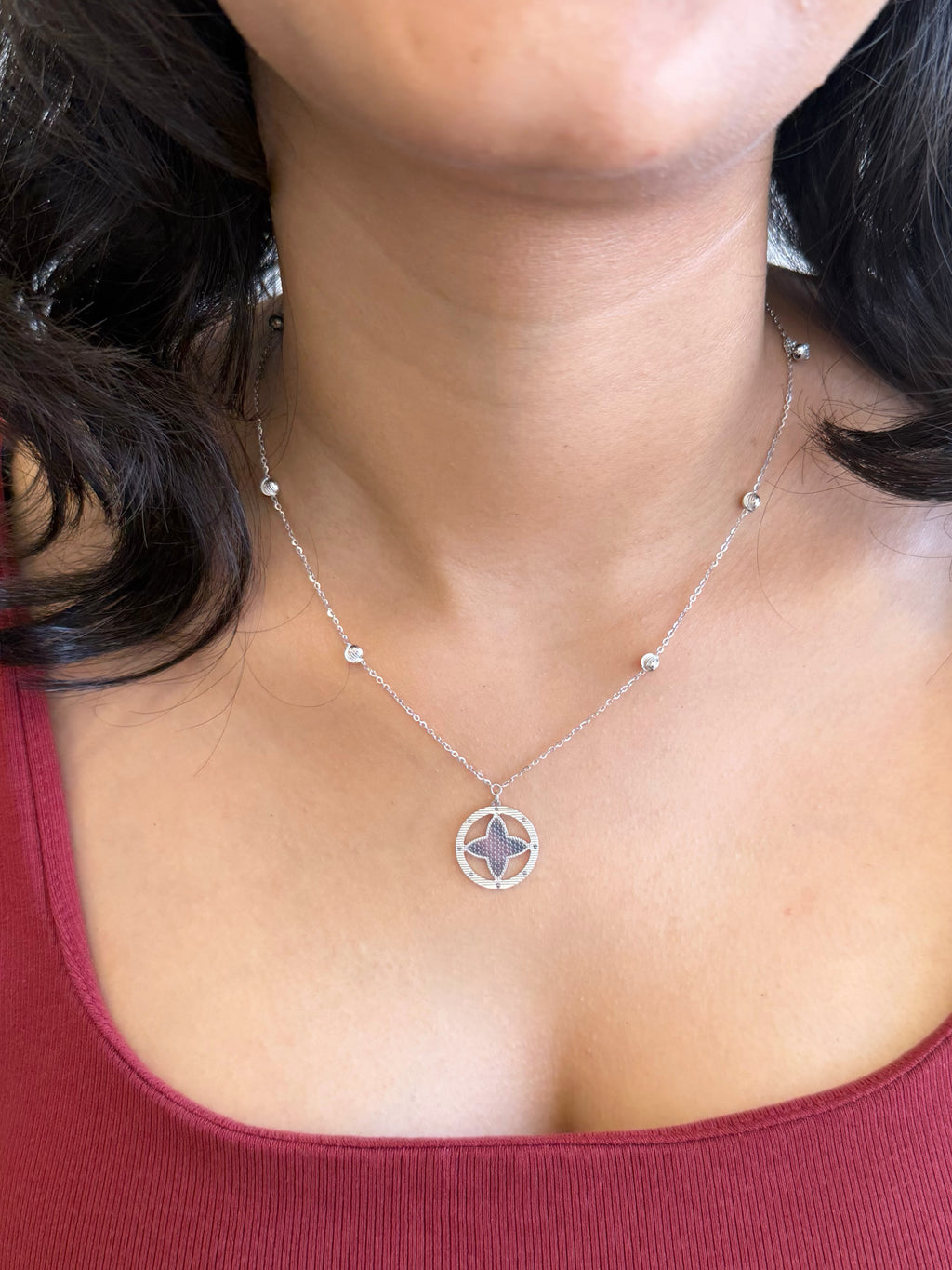 A close-up of a woman wearing a delicate silver necklace with a circular pendant featuring a four-pointed star design, paired with a burgundy ribbed tank top.