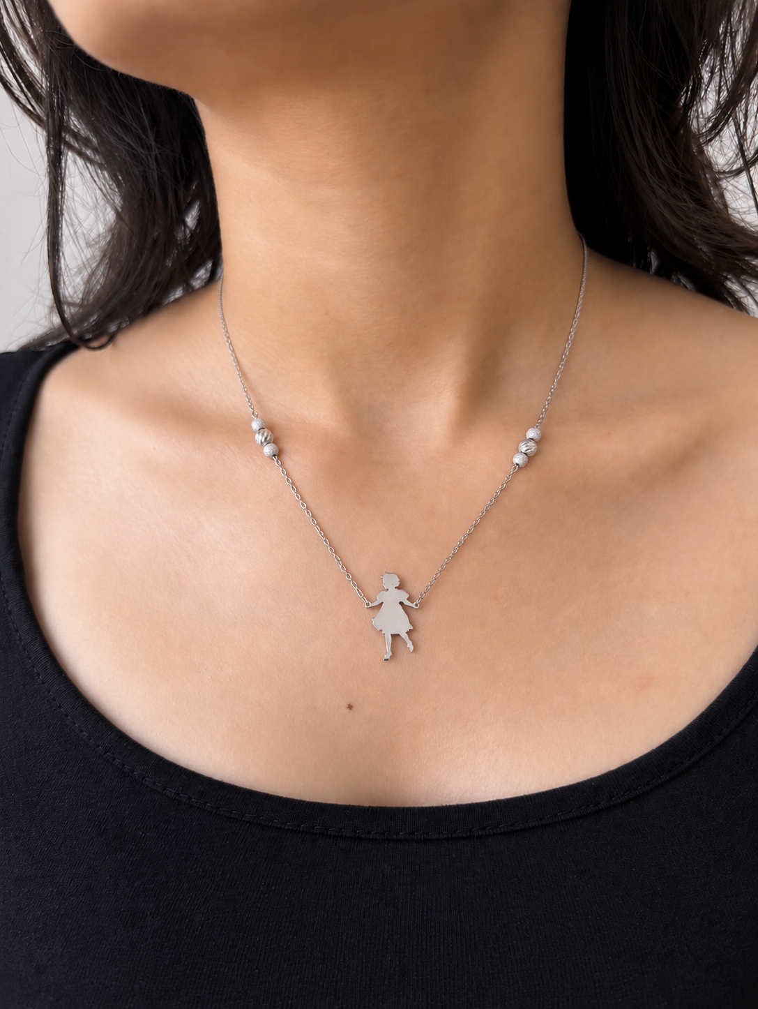 A delicate silver necklace featuring a small silhouette pendant of a dancing girl, suspended from a fine chain with small bead accents on either side, worn by a model who is wearing a black top