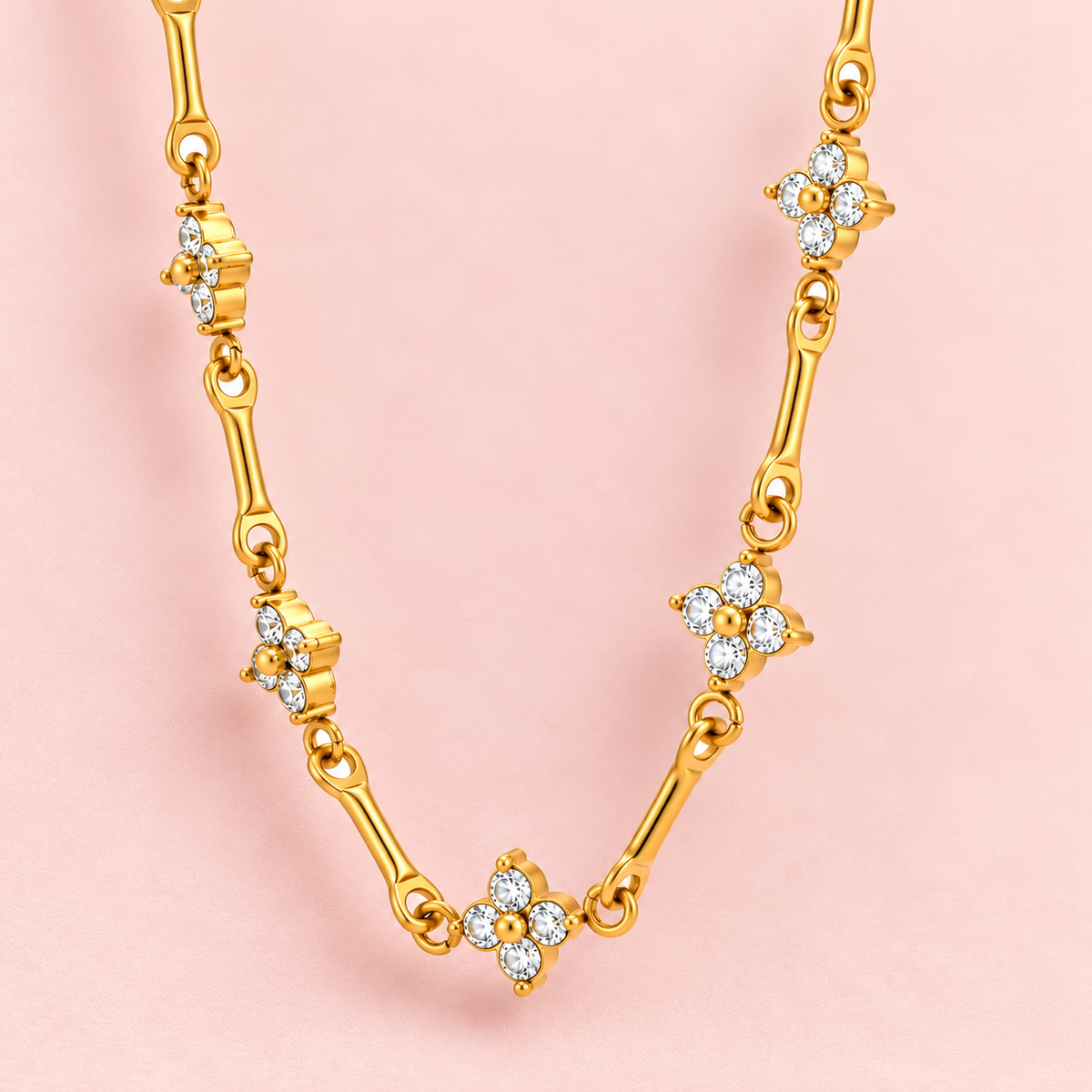Gold-toned floral link necklace featuring four-petal flower motifs with clear sparkling stones and polished gold centres.

Close-up of a delicate gold-tone necklace with floral accents connected by elongated links against a pink background.