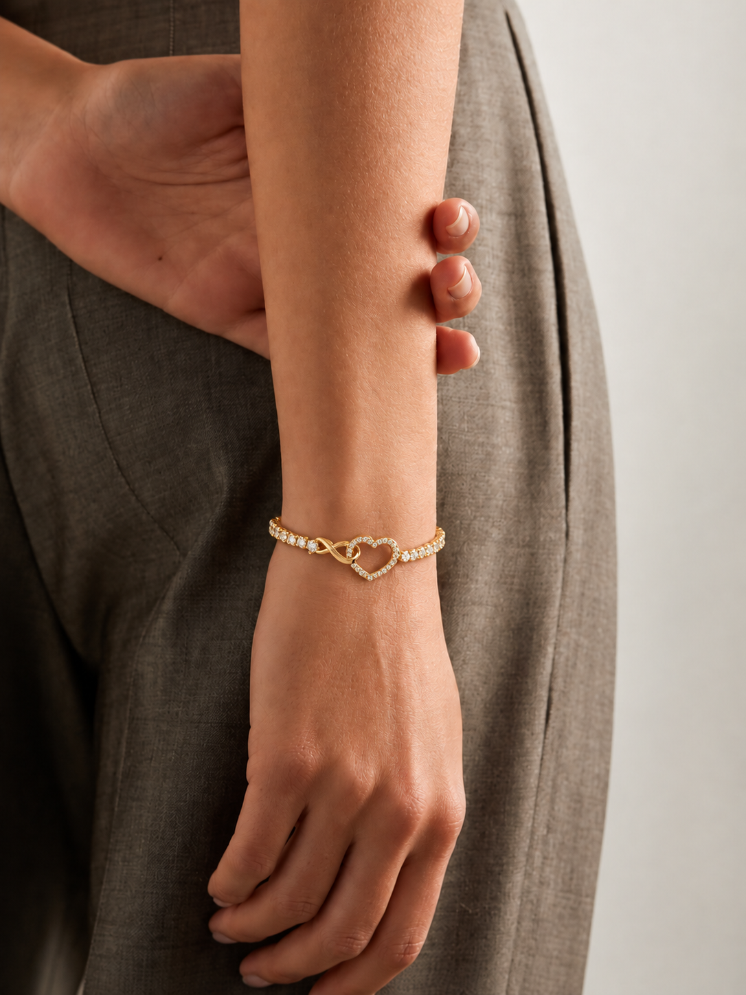 Gold-toned stainless steel bracelet featuring a sparkling open heart intertwined with an infinity motif and white stone accents, adjustable slider chain bracelet with a polished golden finish, worn by a model.