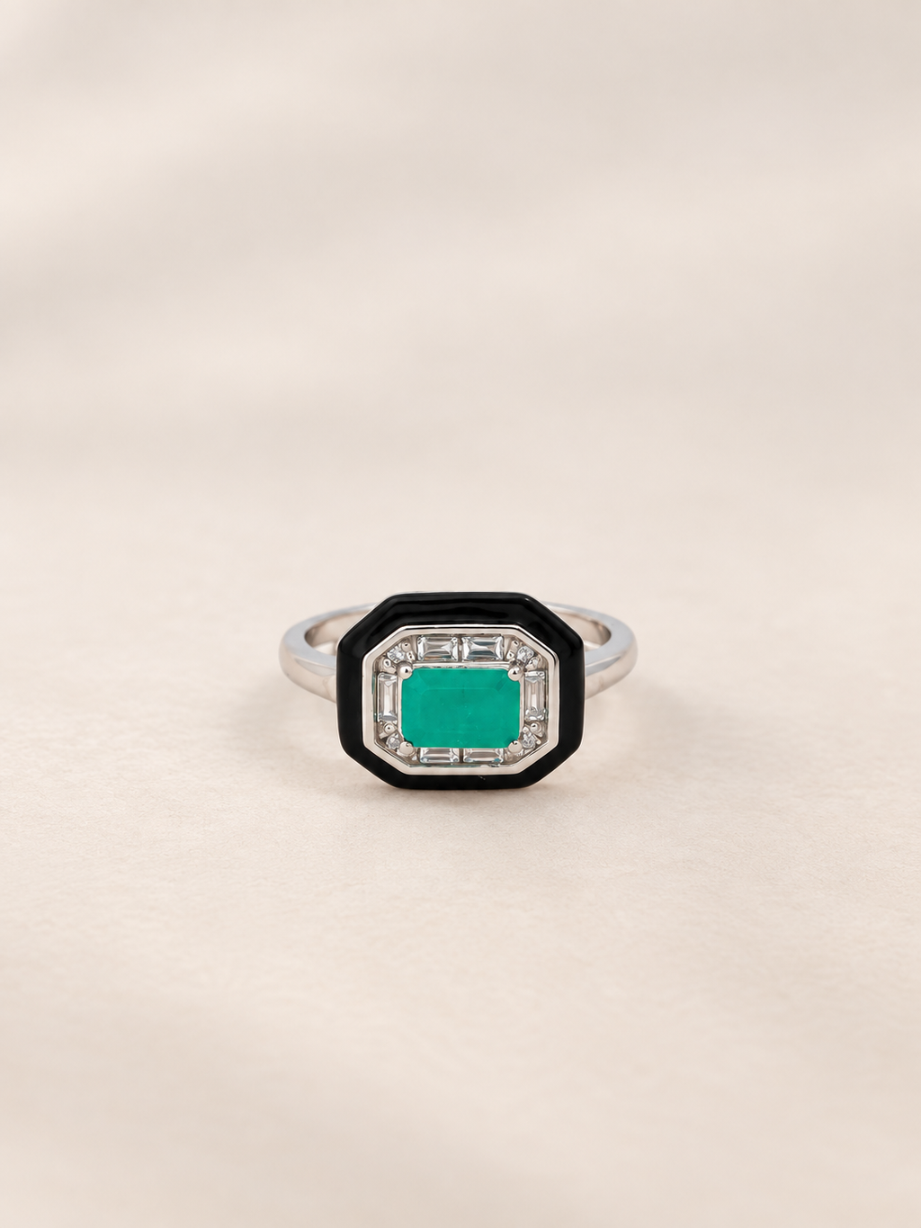 An octagonal ring featuring a vibrant emerald green center stone surrounded by a black and silver geometric border, set on a silver band against a soft beige background.