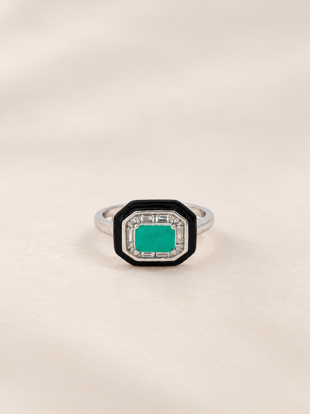 An octagonal ring featuring a vibrant emerald green center stone surrounded by a black and silver geometric border, set on a silver band against a soft beige background.