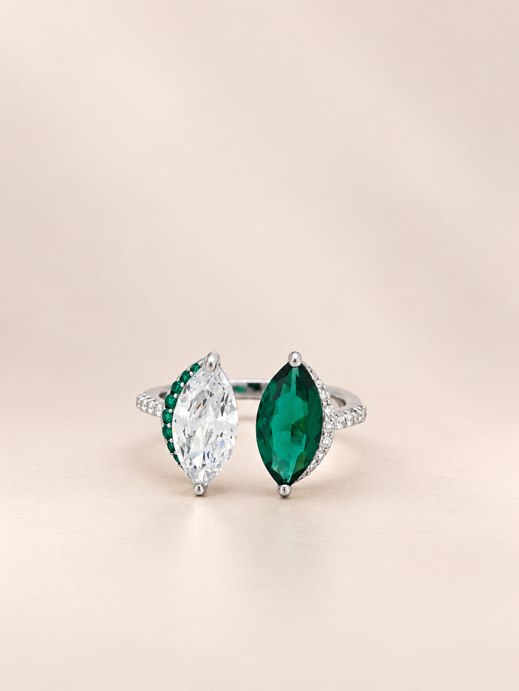 A silver-tone ring featuring two marquise-cut center stones, one clear and one emerald green, with small green and clear accent stones along the shoulders, set against a soft beige background.