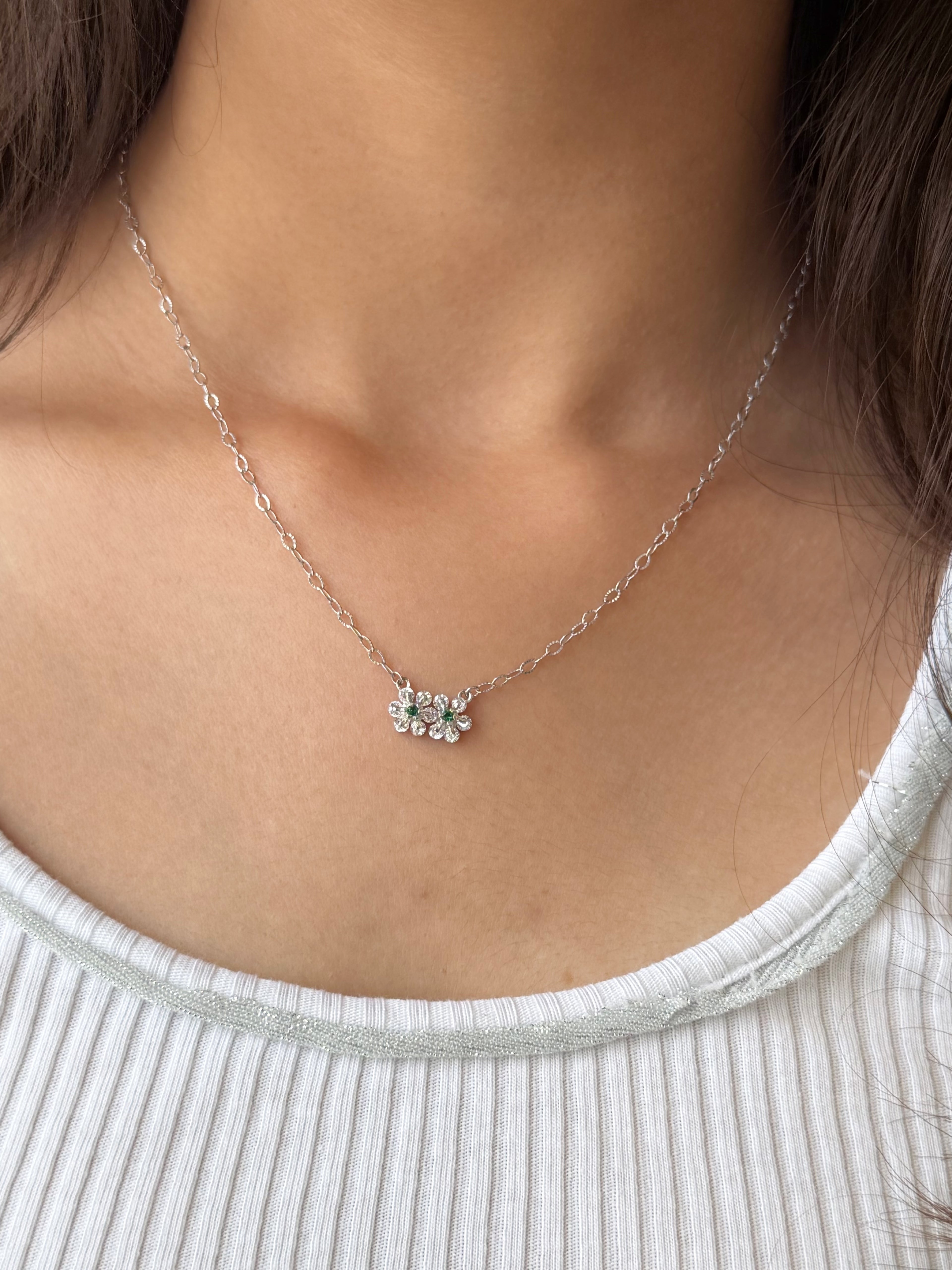 A close-up of a delicate silver chain necklace with a small flower-shaped crystal pendant, worn on a model's neck over a white ribbed top.