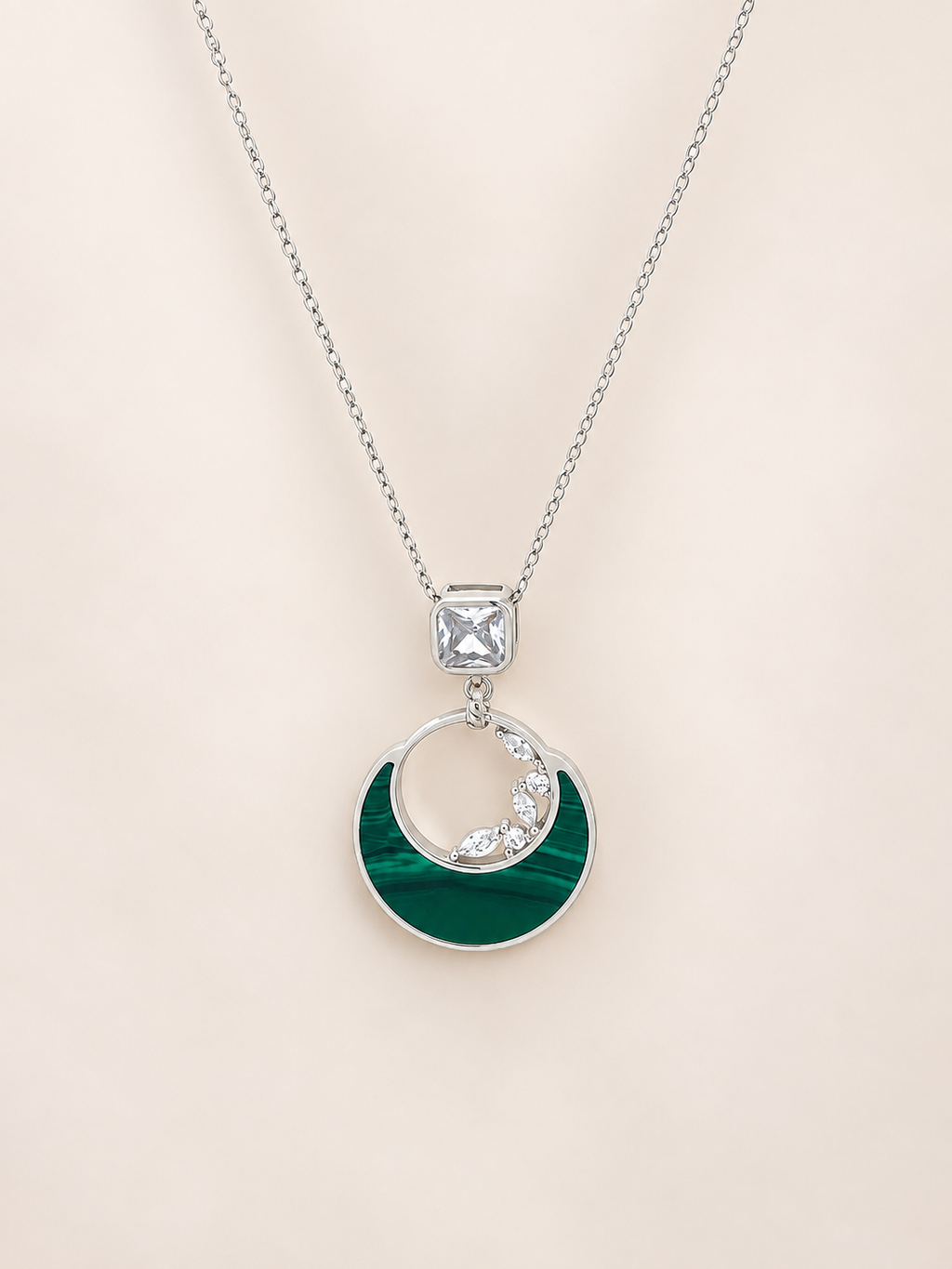 A silver necklace featuring a crescent moon pendant with a dark green inlay and a small row of clear stones along the inner curve, suspended from a square clear stone on a delicate chain, set against a solid pale beige background.
