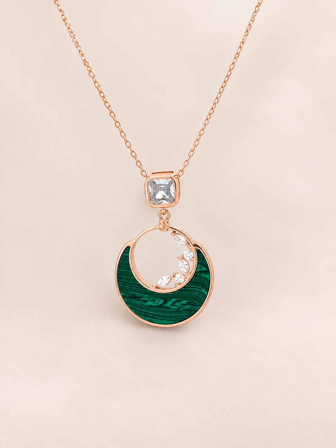 A rose gold pendant necklace featuring a crescent moon design with a textured green inlay and small clear stones, suspended from a delicate rose gold chain on a light beige background.
