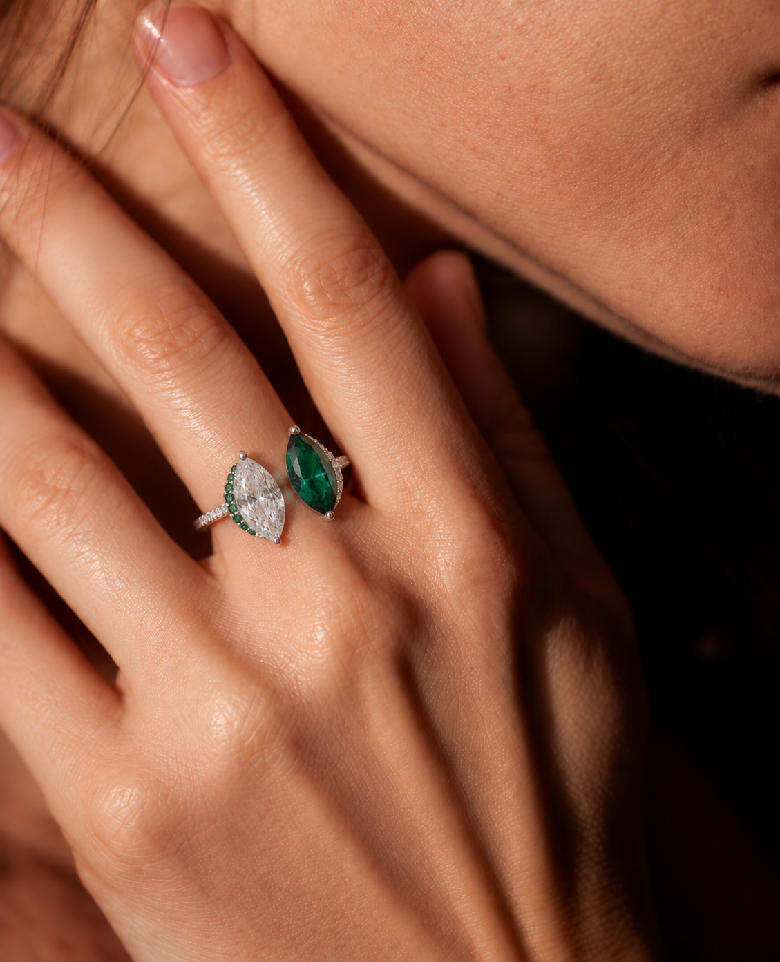 A silver-tone ring featuring two marquise-cut center stones, one clear and one emerald green, with small green and clear accent stones worn by a model.