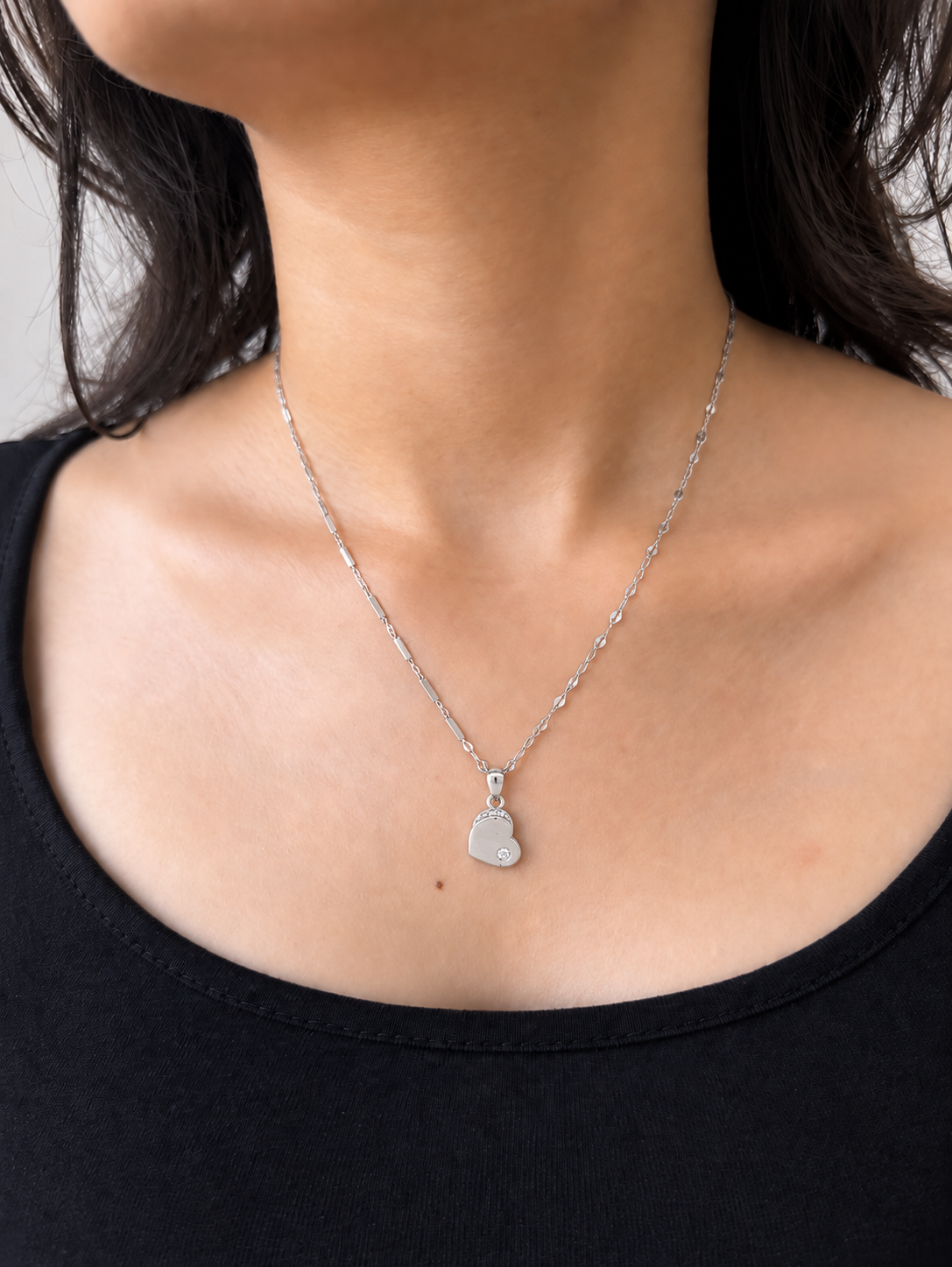 A close-up of a model wearing a delicate silver chain necklace with a small heart-shaped pendant with a small diamond inside.