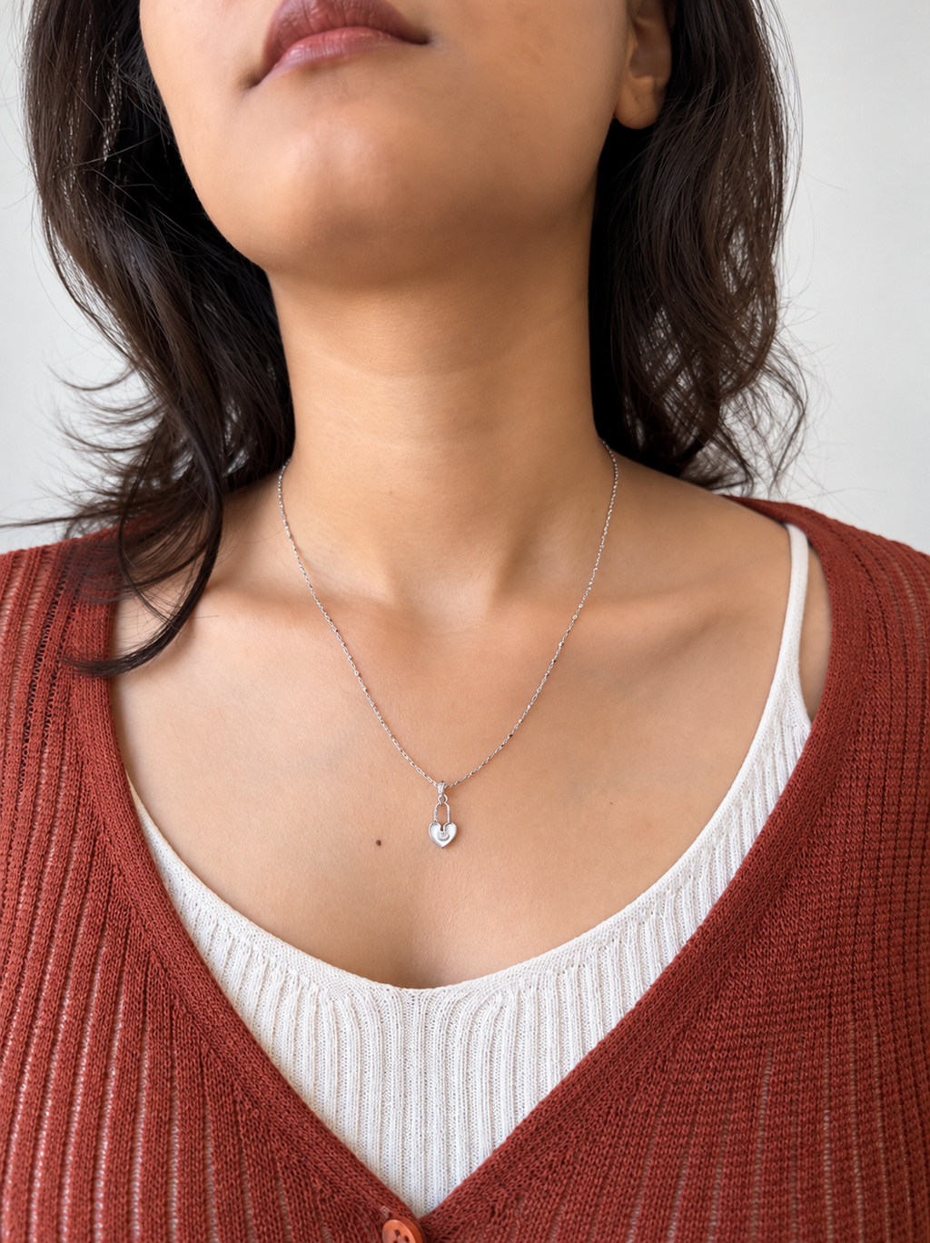 A close-up of a woman wearing a delicate silver chain necklace with a small heart-shaped pendant, styled with a white ribbed tank top and a rust-colored knit cardigan.