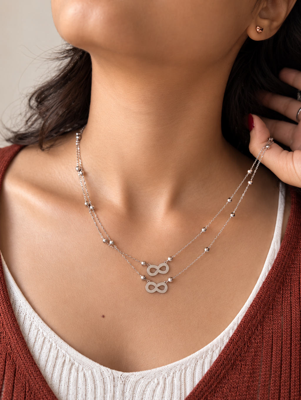 A close-up of a model wearing a delicate silver double-strand necklace with small bead accents and two infinity symbols at the center, styled with a white ribbed top and a rust-colored cardigan.