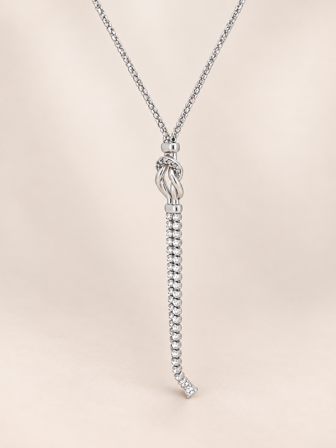 A silver-toned necklace featuring a delicate chain and a pendant with a knotted top and a long, beaded drop, displayed against a soft beige background.