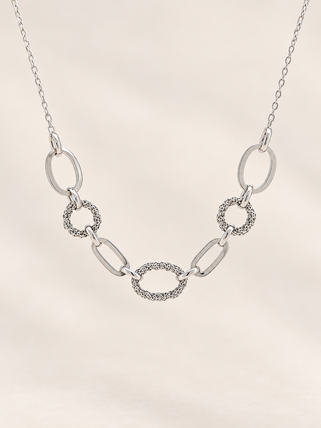 A silver link chain necklace featuring a central oval link with a textured, pavé-like border, flanked by polished oval and textured circular links, displayed on a soft beige background.