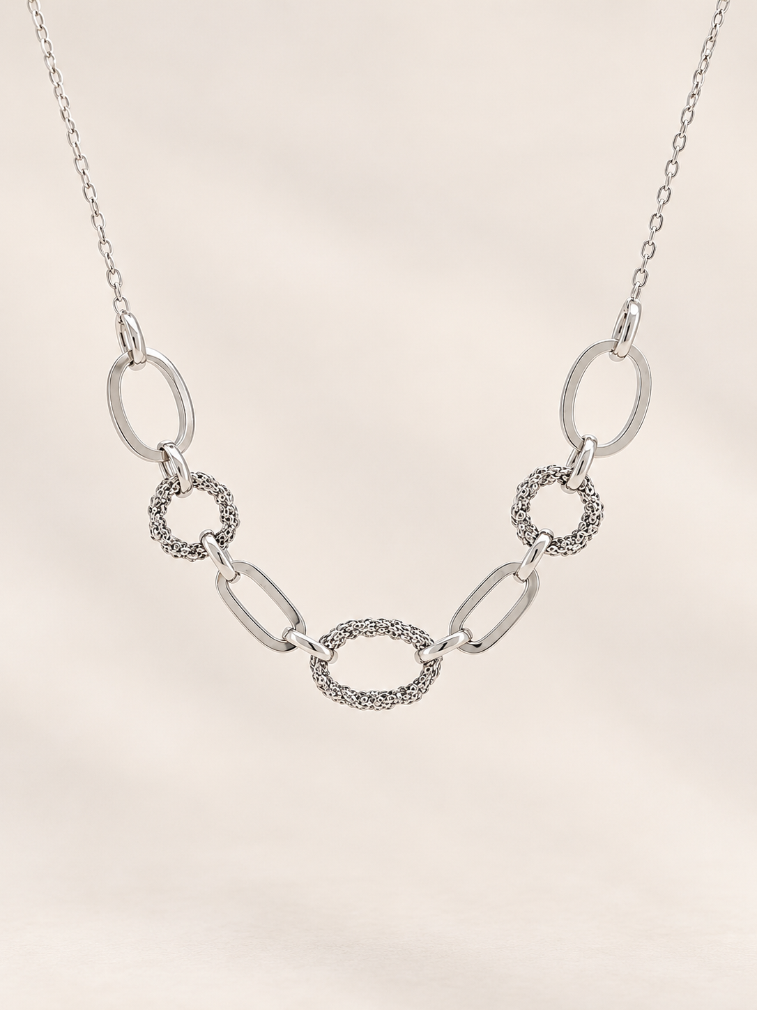 A silver link chain necklace featuring a central oval link with a textured, pavé-like border, flanked by polished oval and textured circular links, displayed on a soft beige background.