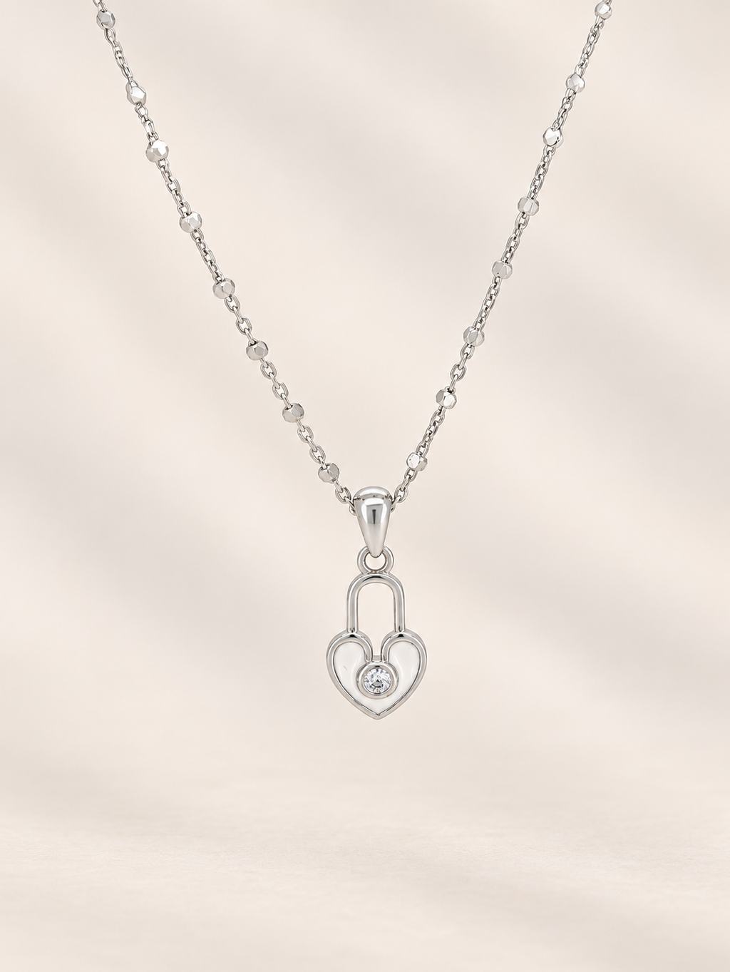A silver heart-shaped pendant necklace with a small clear gemstone in the center, suspended from a delicate beaded chain against a soft beige background.