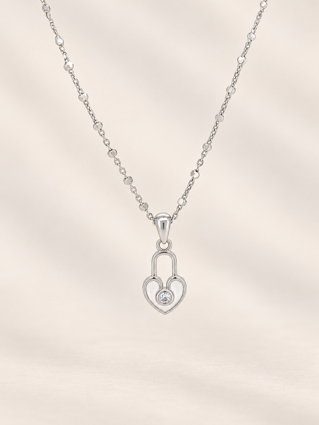 A silver heart-shaped pendant necklace with a small clear gemstone in the center, suspended from a delicate beaded chain against a soft beige background.