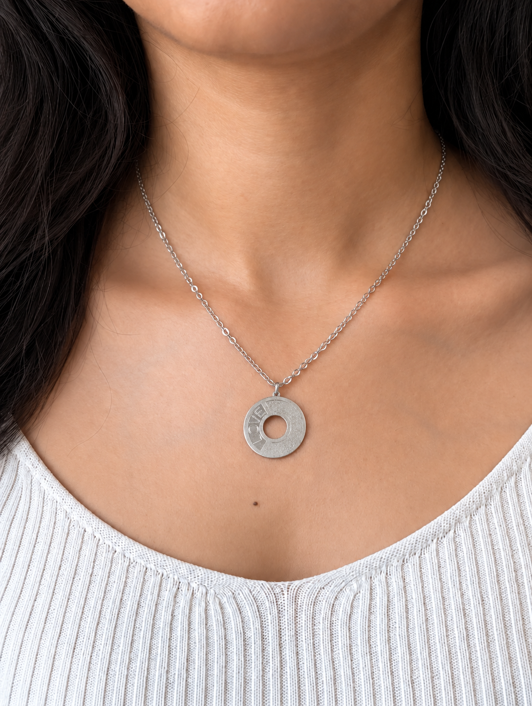 A silver-tone chain necklace with a textured circular disc pendant engraved with the word 'LOVE', worn by a model wearing a white top.