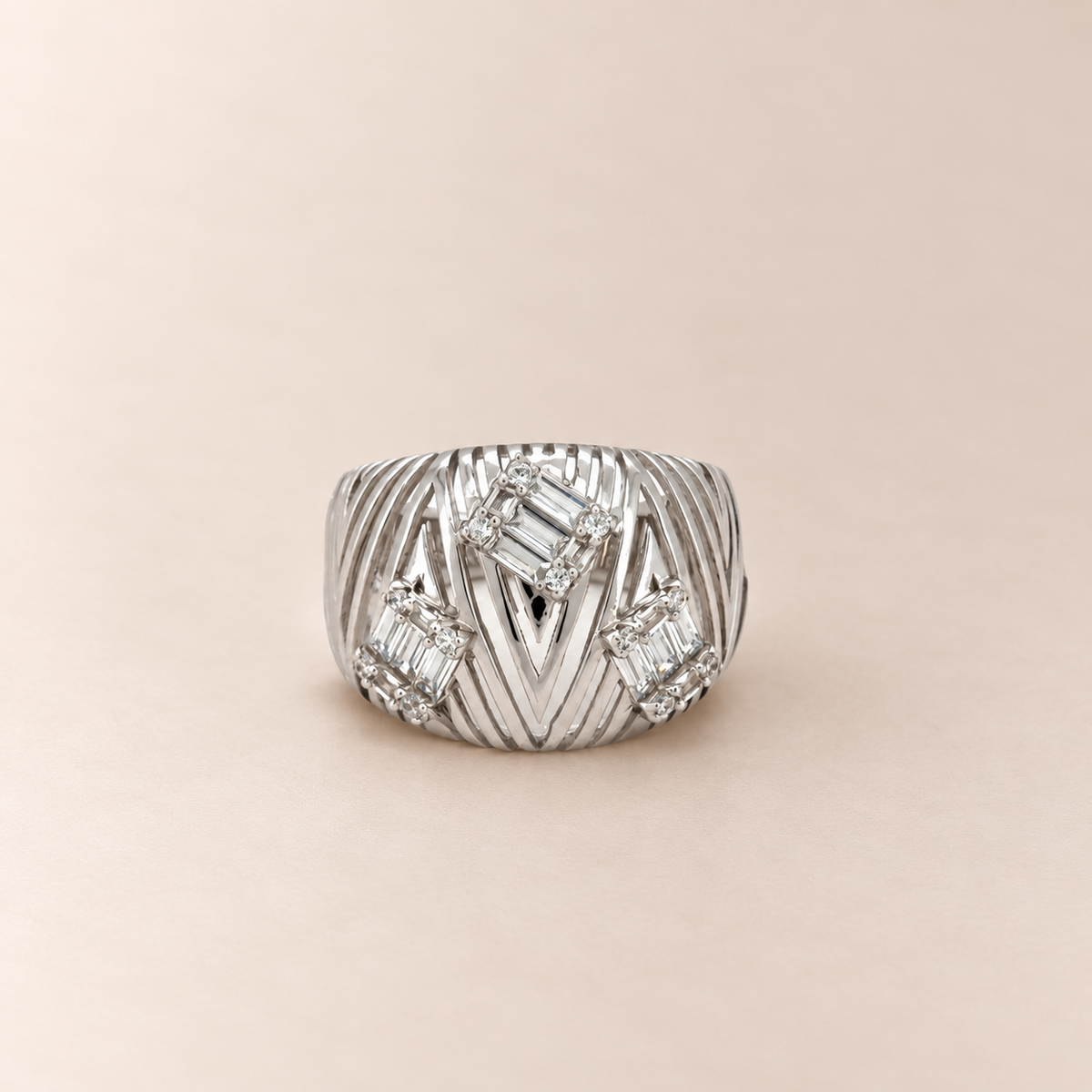 A silver-toned statement ring with a textured, ribbed band and baguette-cut clear stones, set against a solid pale pink background.