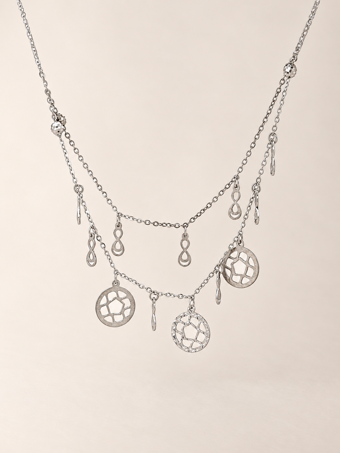 A silver-toned multi-layer necklace featuring delicate chains, teardrop charms, and circular filigree pendants, displayed against a solid light beige background.